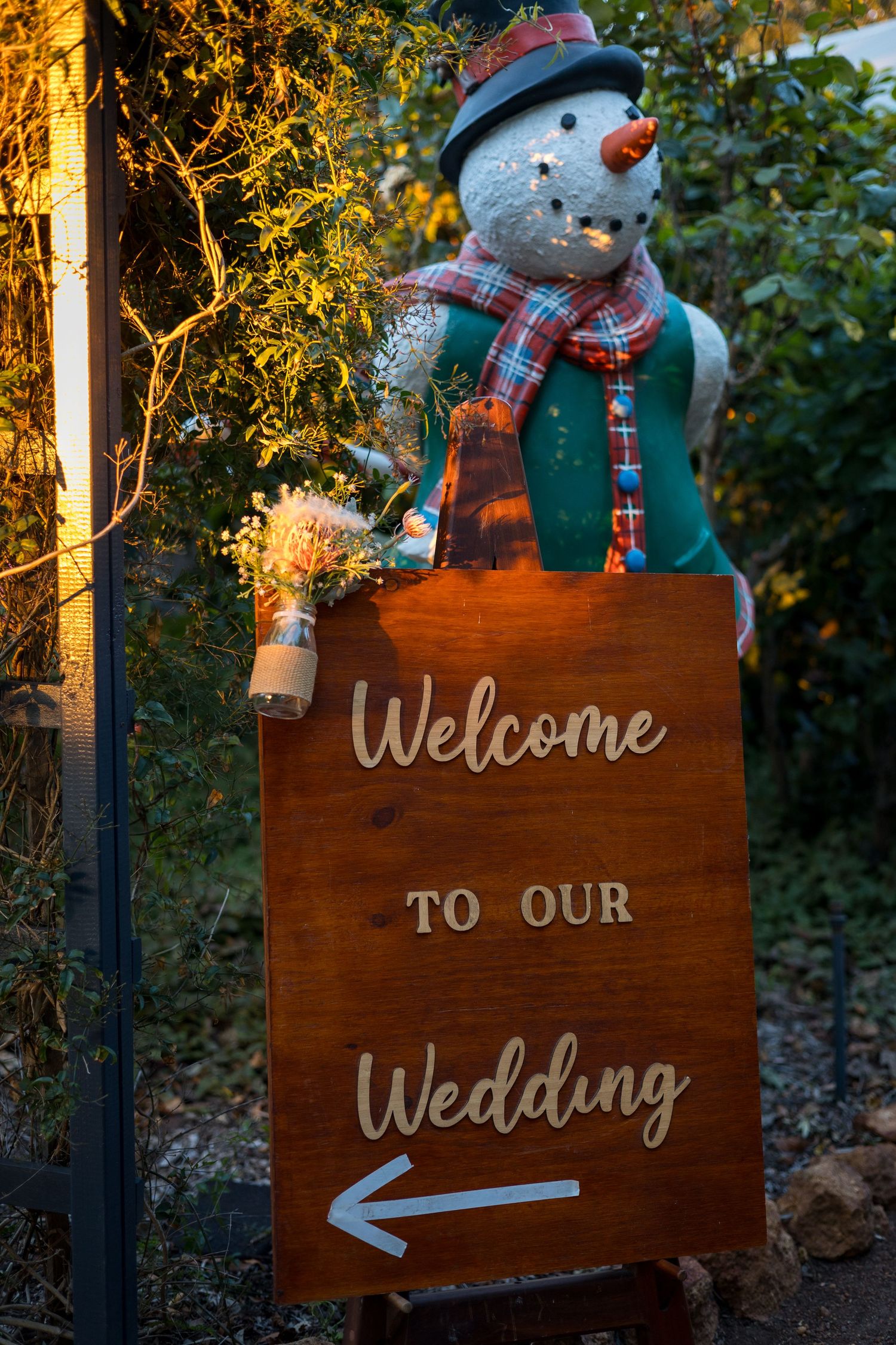Rustic wooden wedding welcome sign decorated with a snowman figure and plaid scarf in an outdoor garden setting.