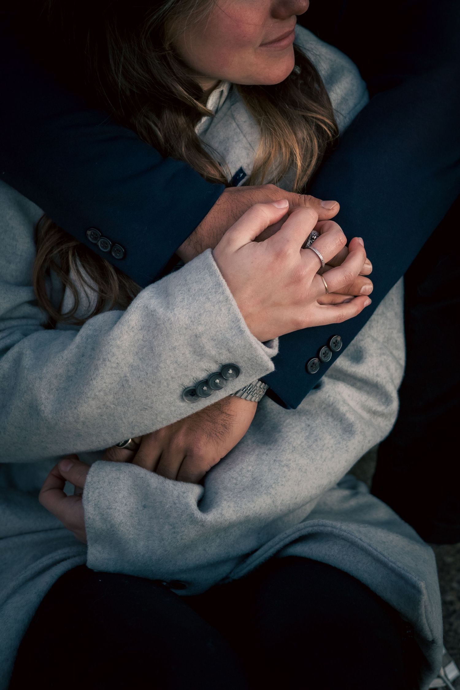 Close-up of couple embracing, hands intertwined, showcasing wedding rings against elegant grey and navy attire.