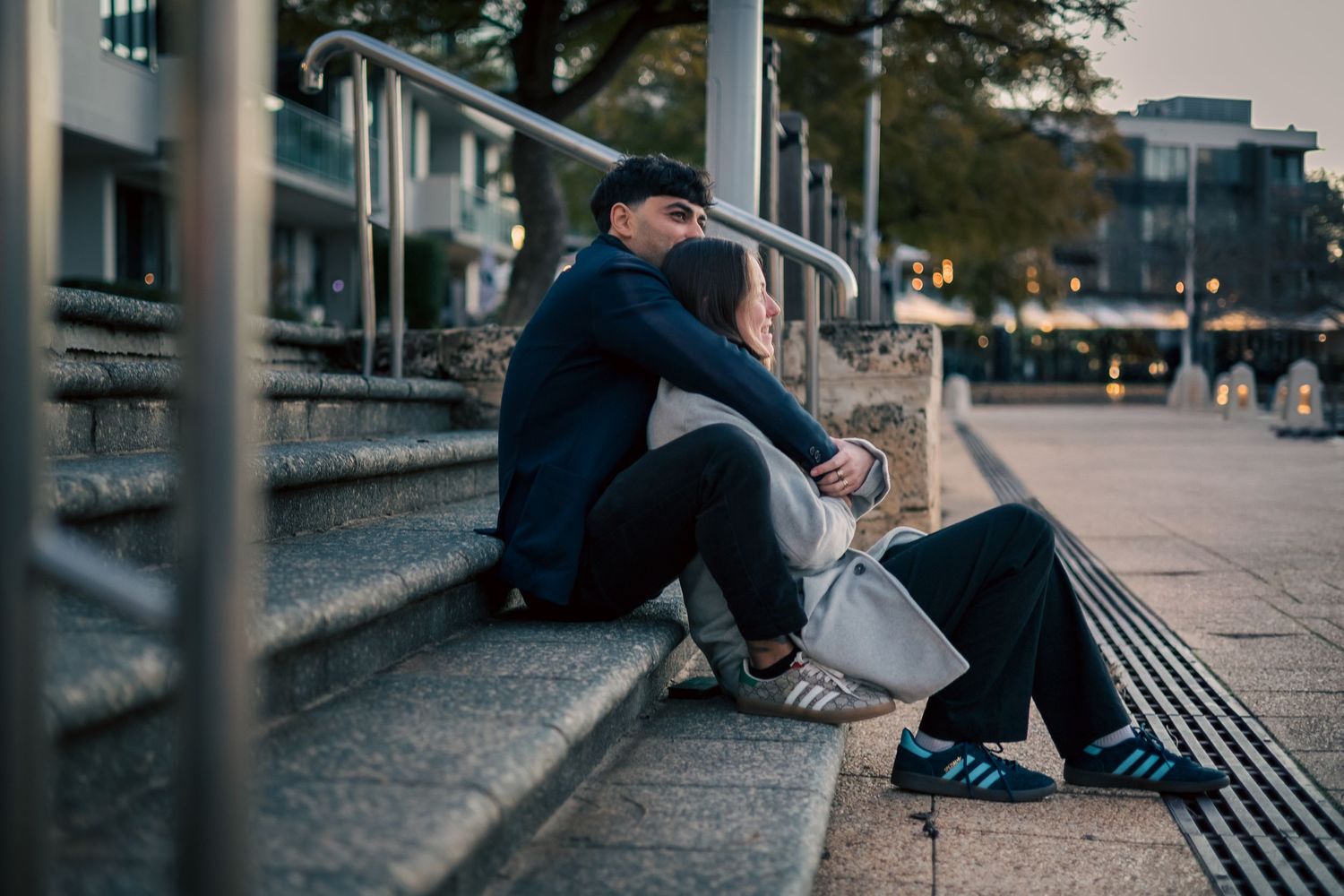 Couple sitting closely on urban steps at dusk, sharing a tender embrace in a city setting.