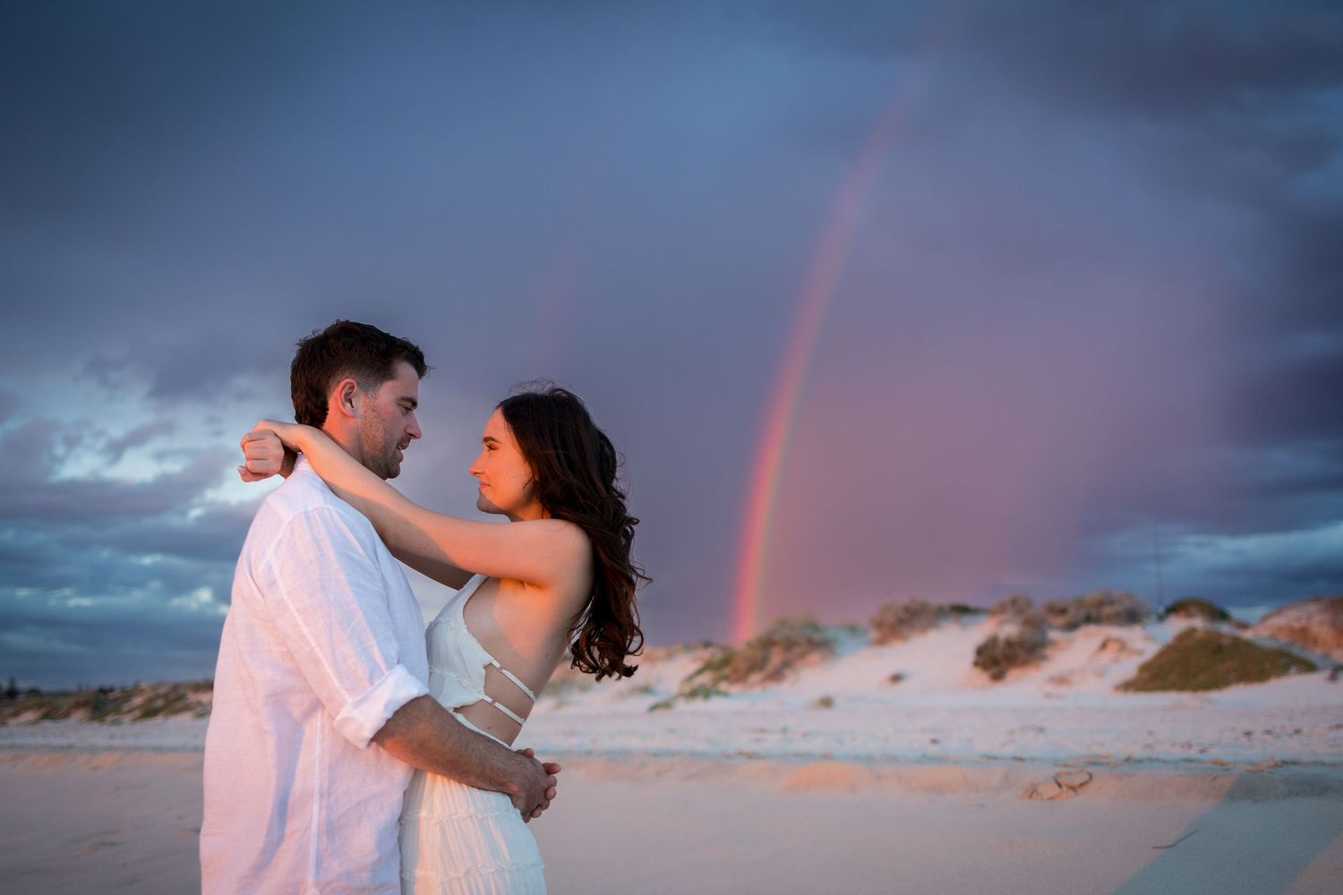 Couple embracing on a beach at sunset with a vibrant rainbow over sand dunes in the background.