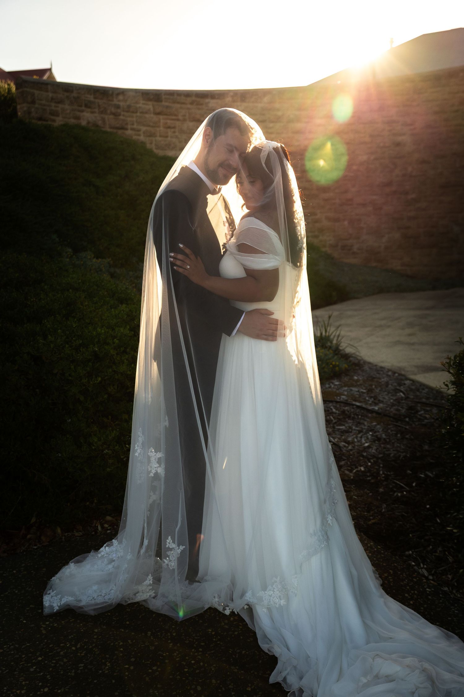 Newlyweds embrace under a flowing cathedral veil with golden sunset light behind them outdoors.