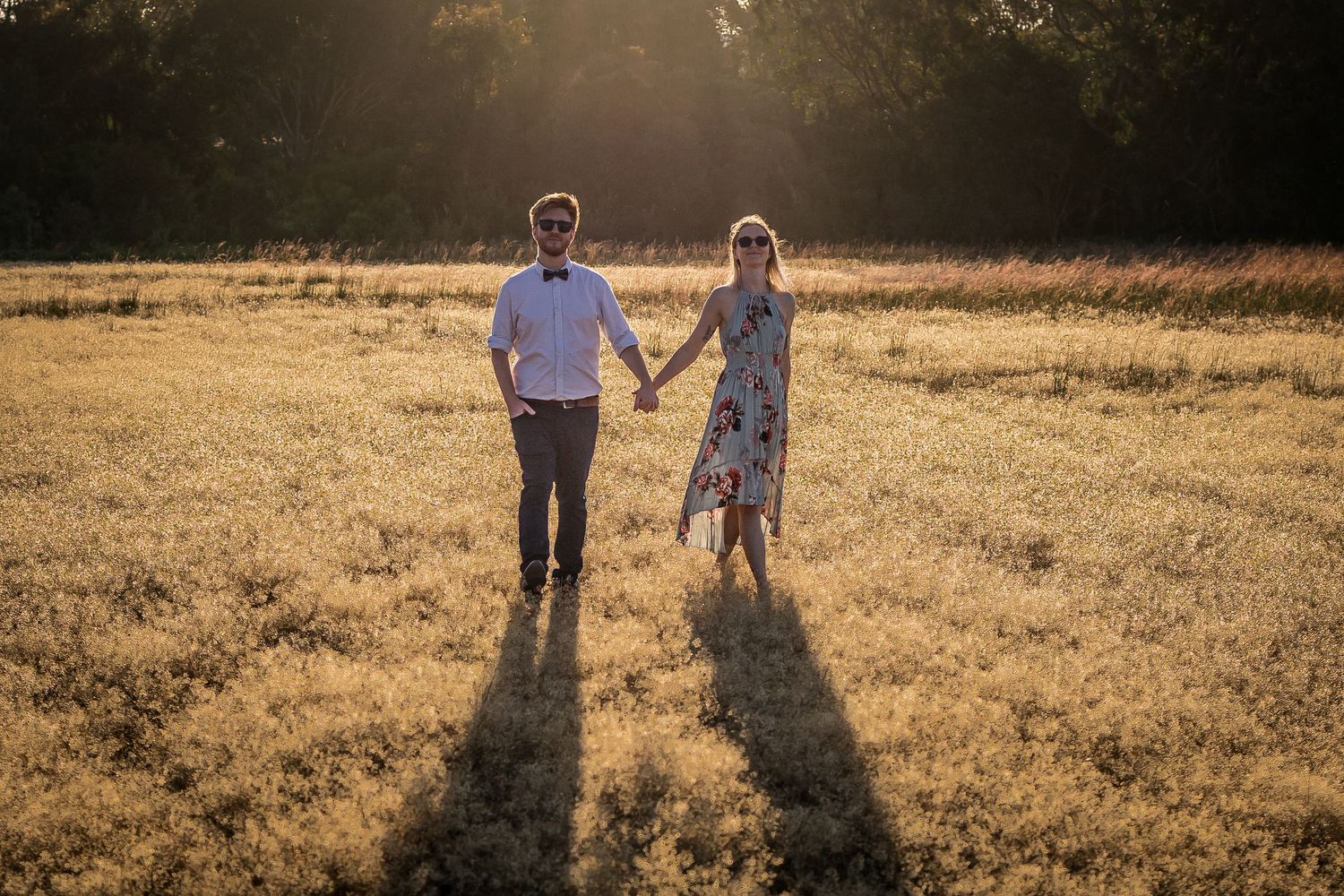 Couple holding hands walking through golden field at sunset casting long shadows.