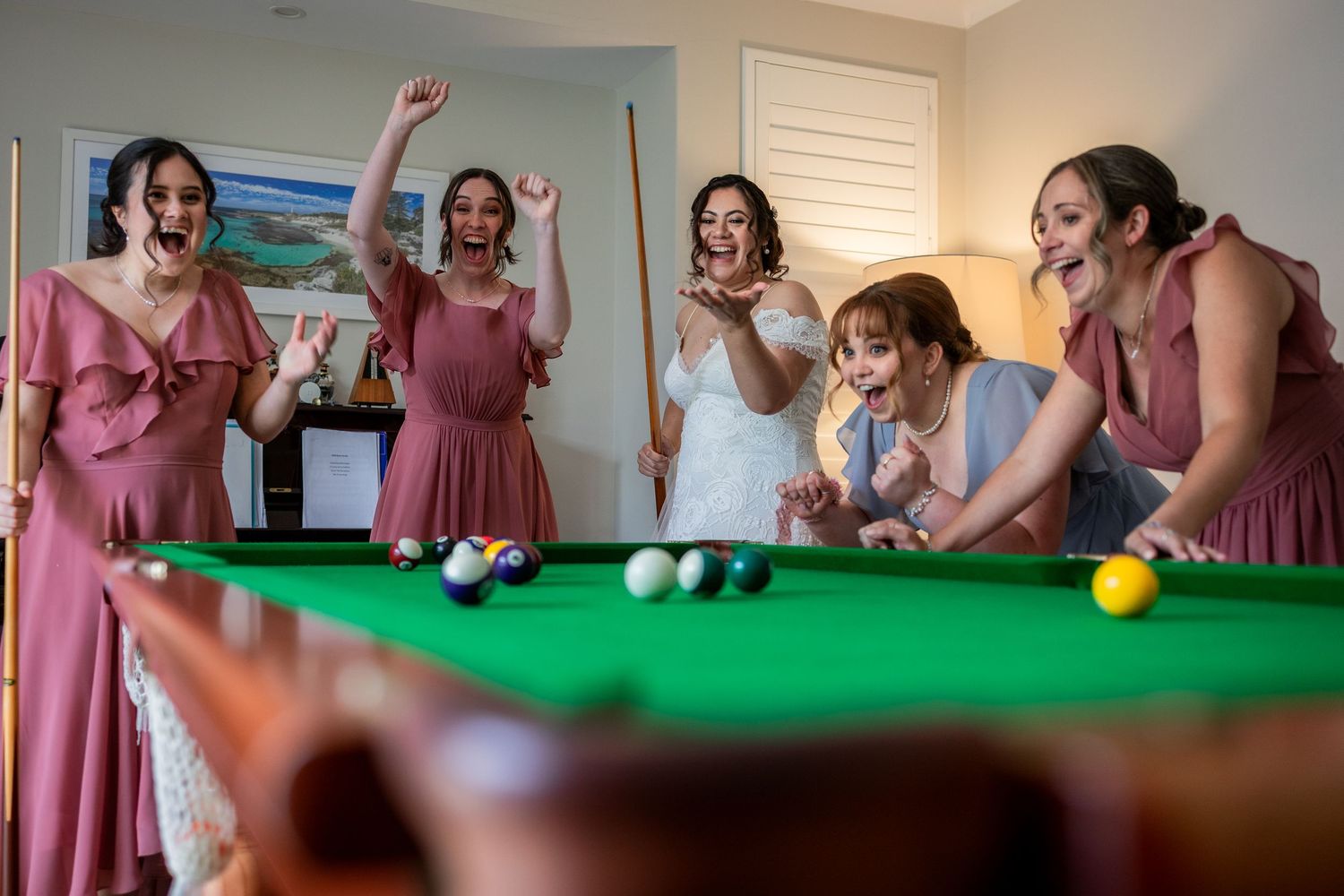 Bride and bridesmaids in pink dresses celebrating around a pool table.