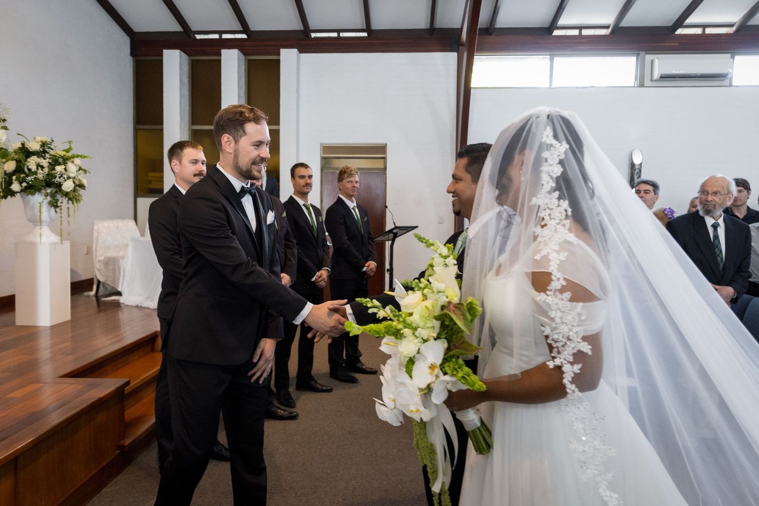 Groom shaking hands with bride holding white orchids at elegant indoor wedding ceremony with guests in background.
