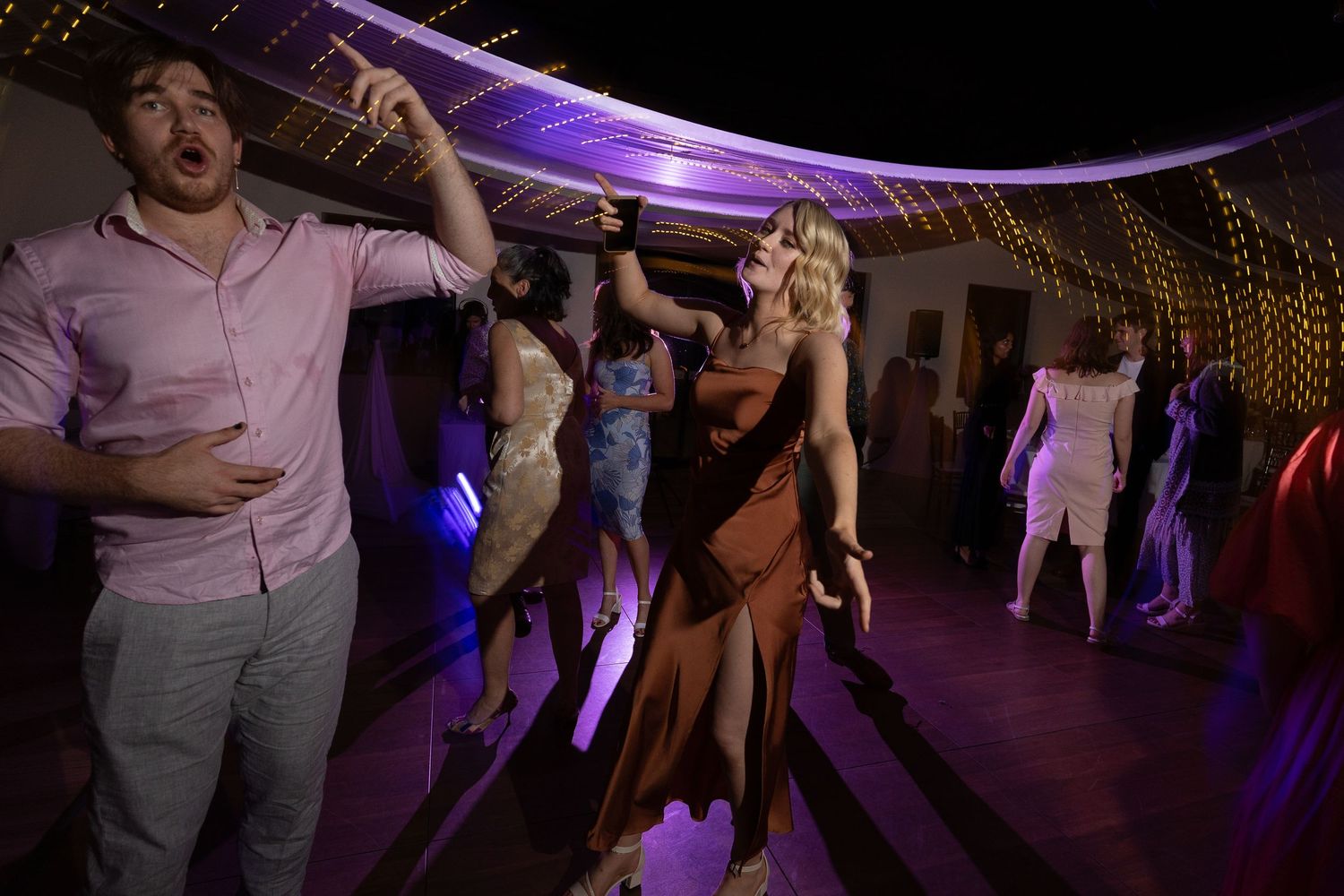 Party guests dancing enthusiastically under purple mood lighting at a wedding reception.