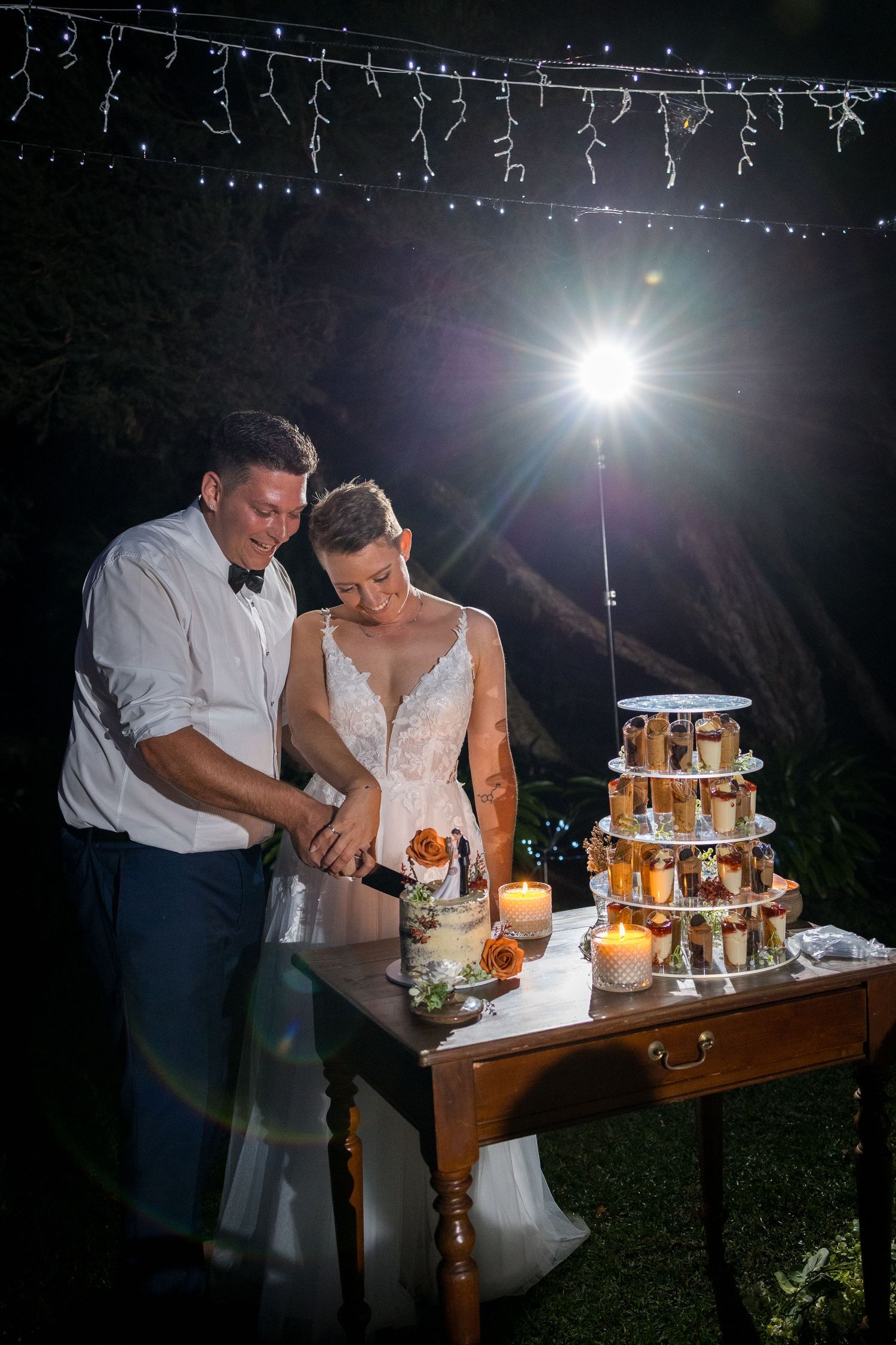 A couple cuts their multi-tiered wedding cake at an outdoor evening reception under twinkling lights.