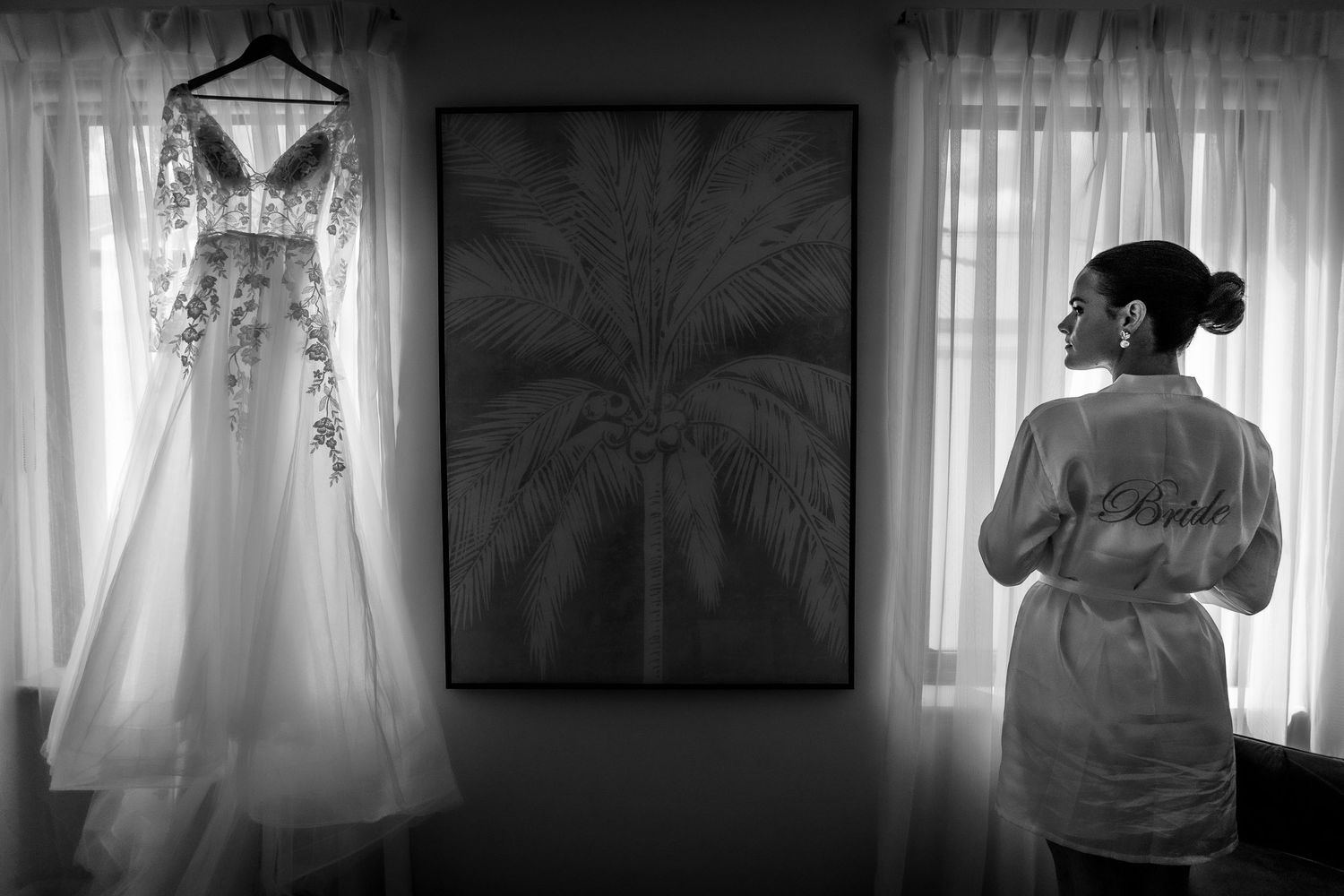 Bride in robe gazes at her floral wedding gown hanging by a window in a black and white photo.