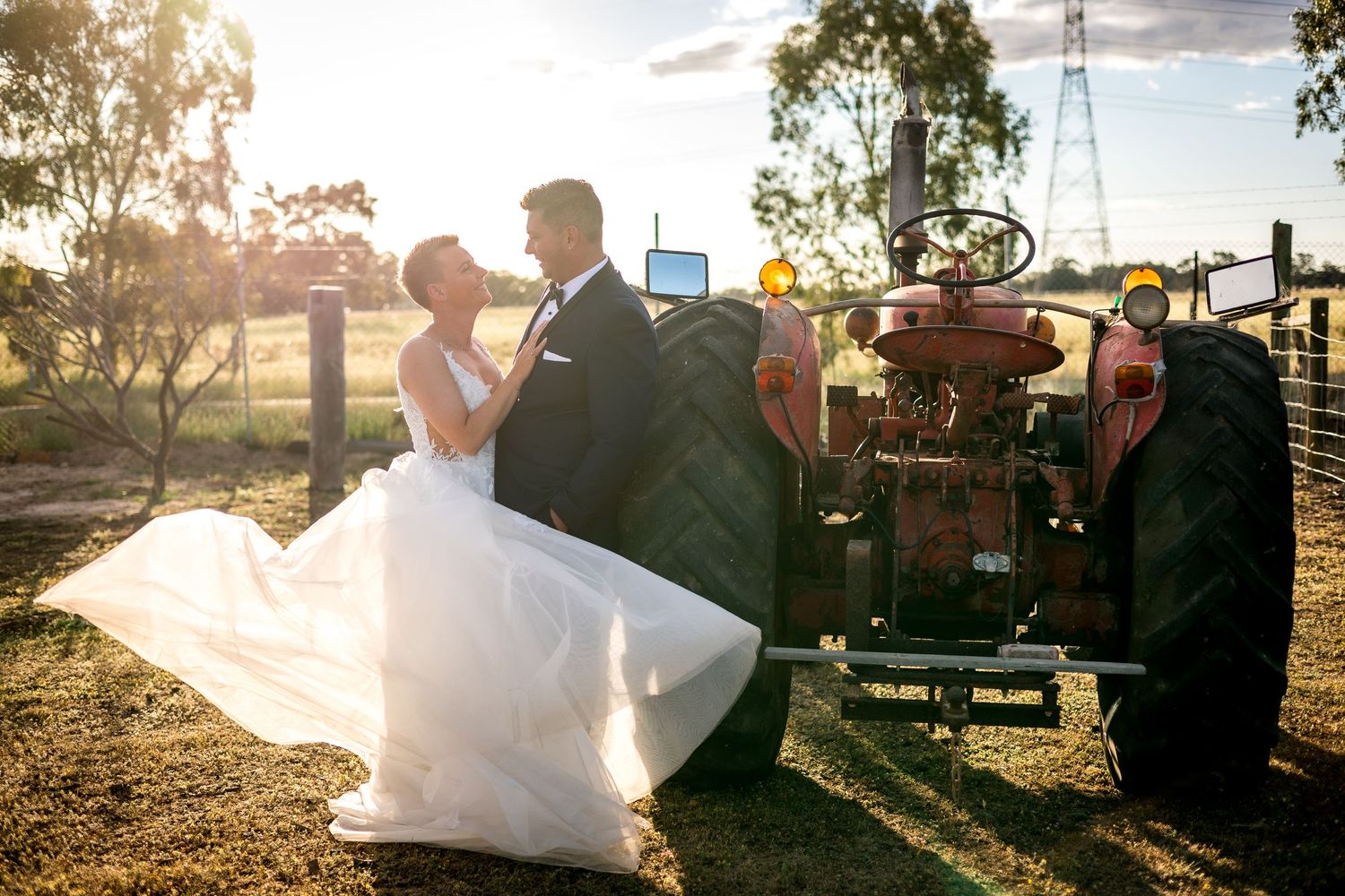 Bride and groom share romantic kiss beside vintage red tractor at golden hour on rustic farm wedding venue.