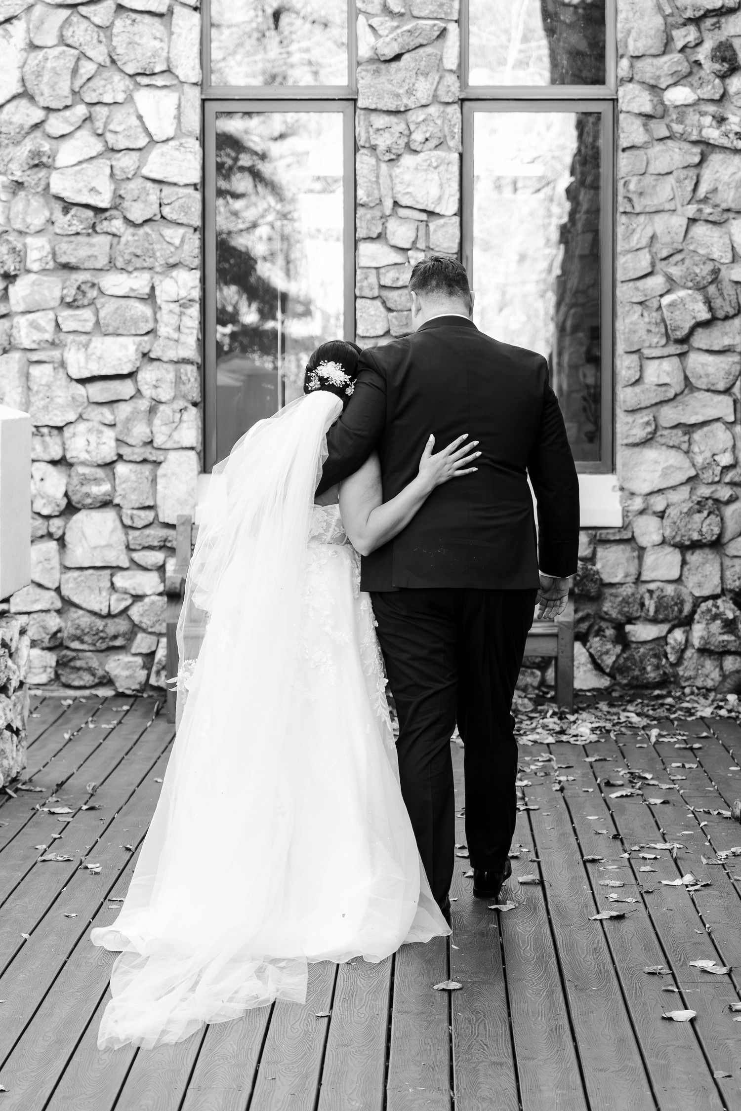Bride and groom walk arm in arm toward stone building in elegant black and white wedding photo.