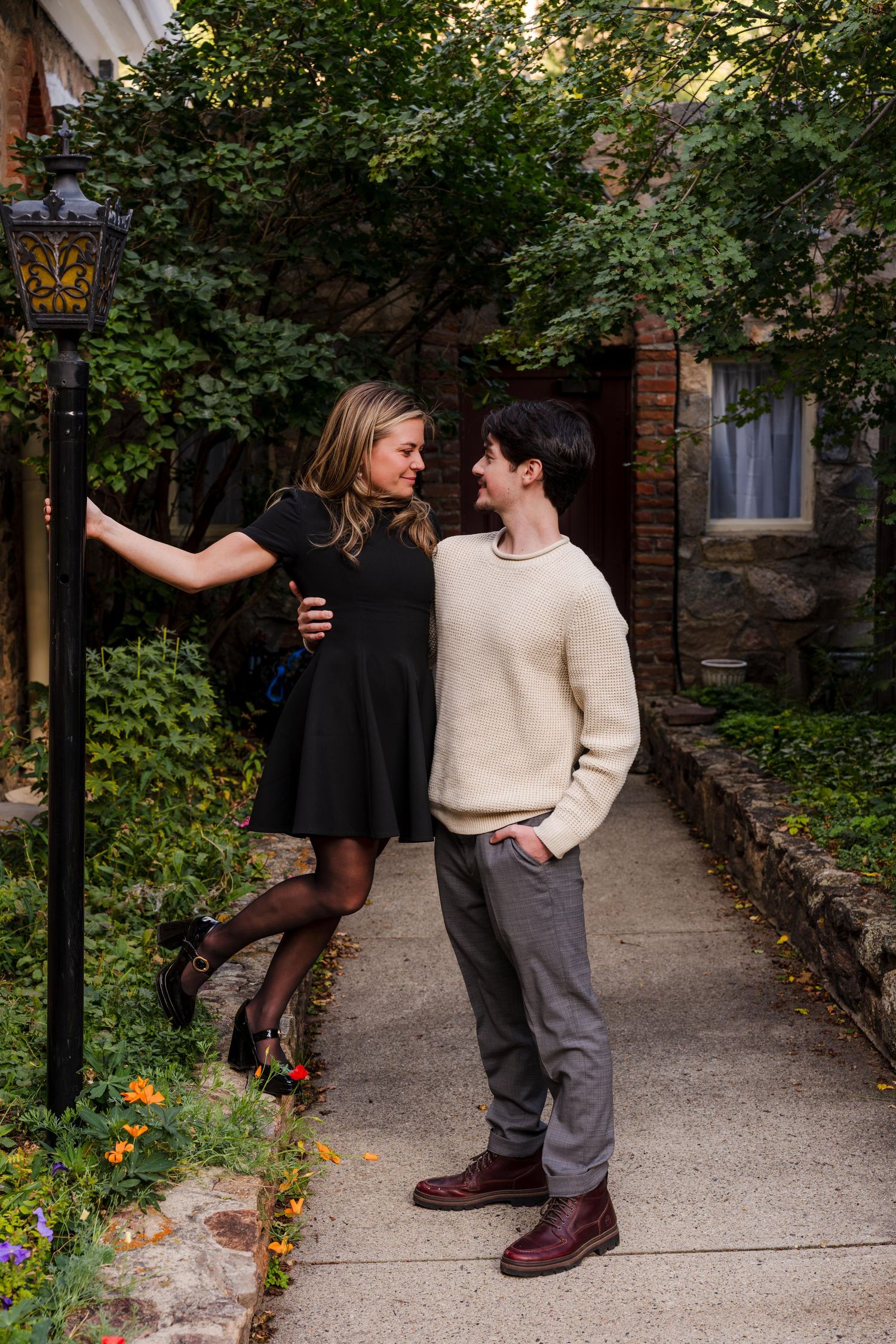 Couple sharing a romantic moment on a garden pathway, woman in black dress leaning on lamppost, man in cream sweater.