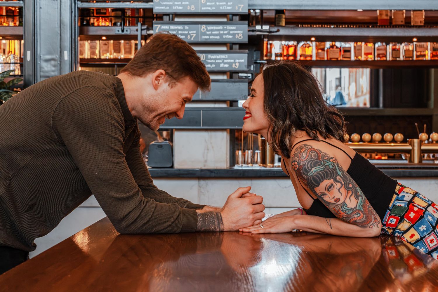 Man and tattooed woman lean across bar counter smiling at each other in trendy bar setting.