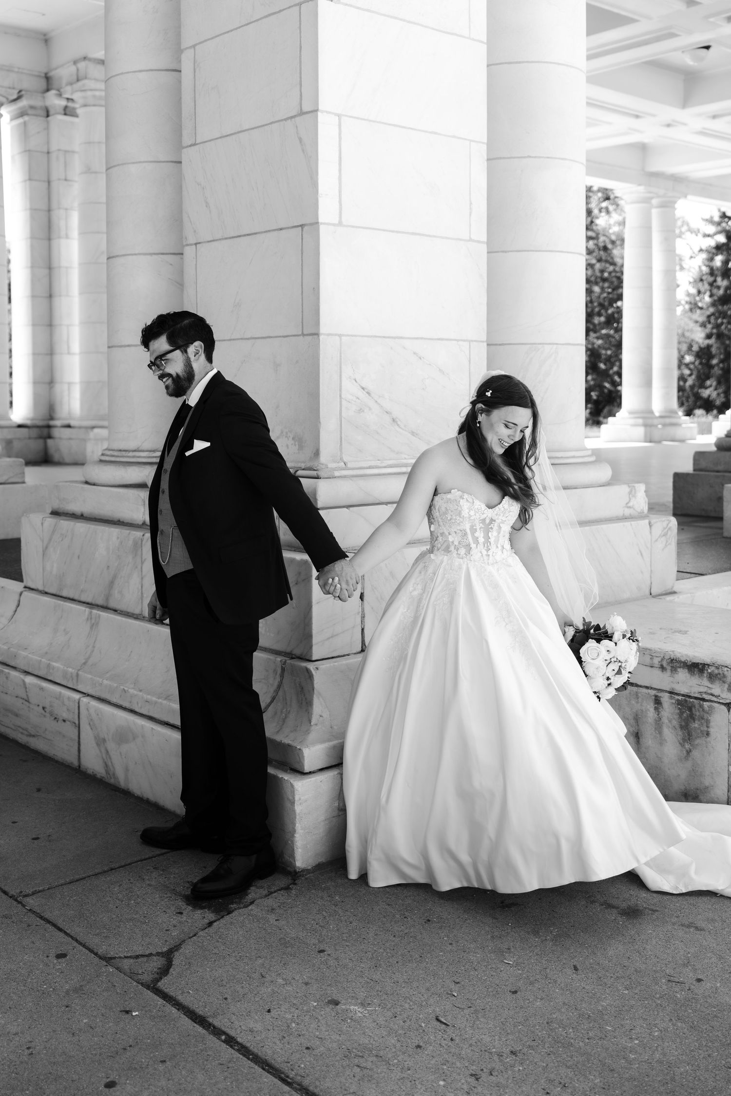 Black and white photo of a bride and groom holding hands near marble columns on their wedding day.