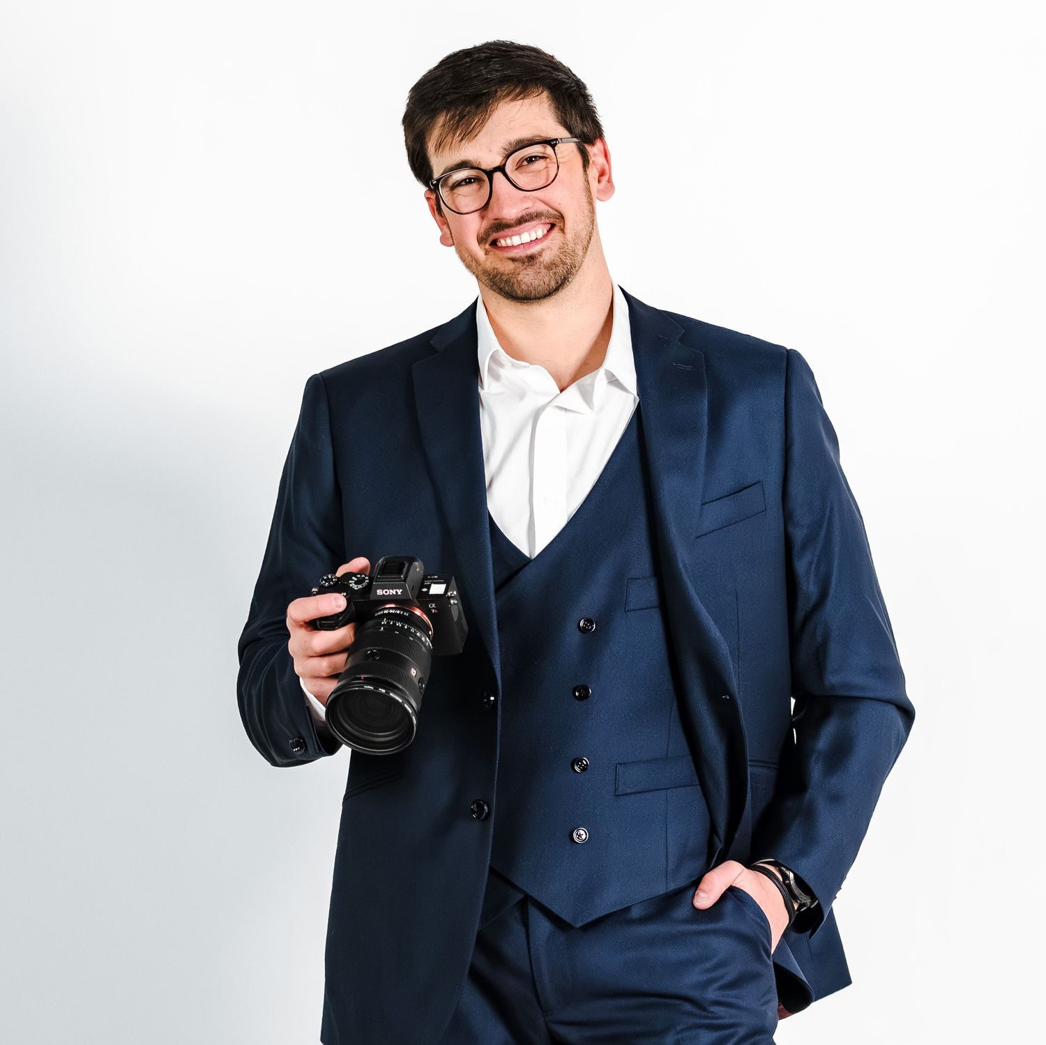 Smiling man in navy blue three-piece suit holding a DSLR camera, posing against a white background.