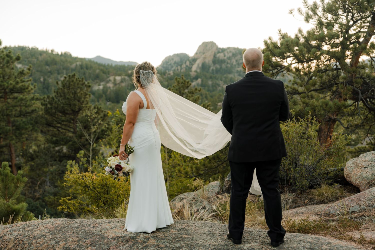 Bride and groom stand on rocky overlook with mountain views, bride's veil flowing in wind, Colorado wedding.