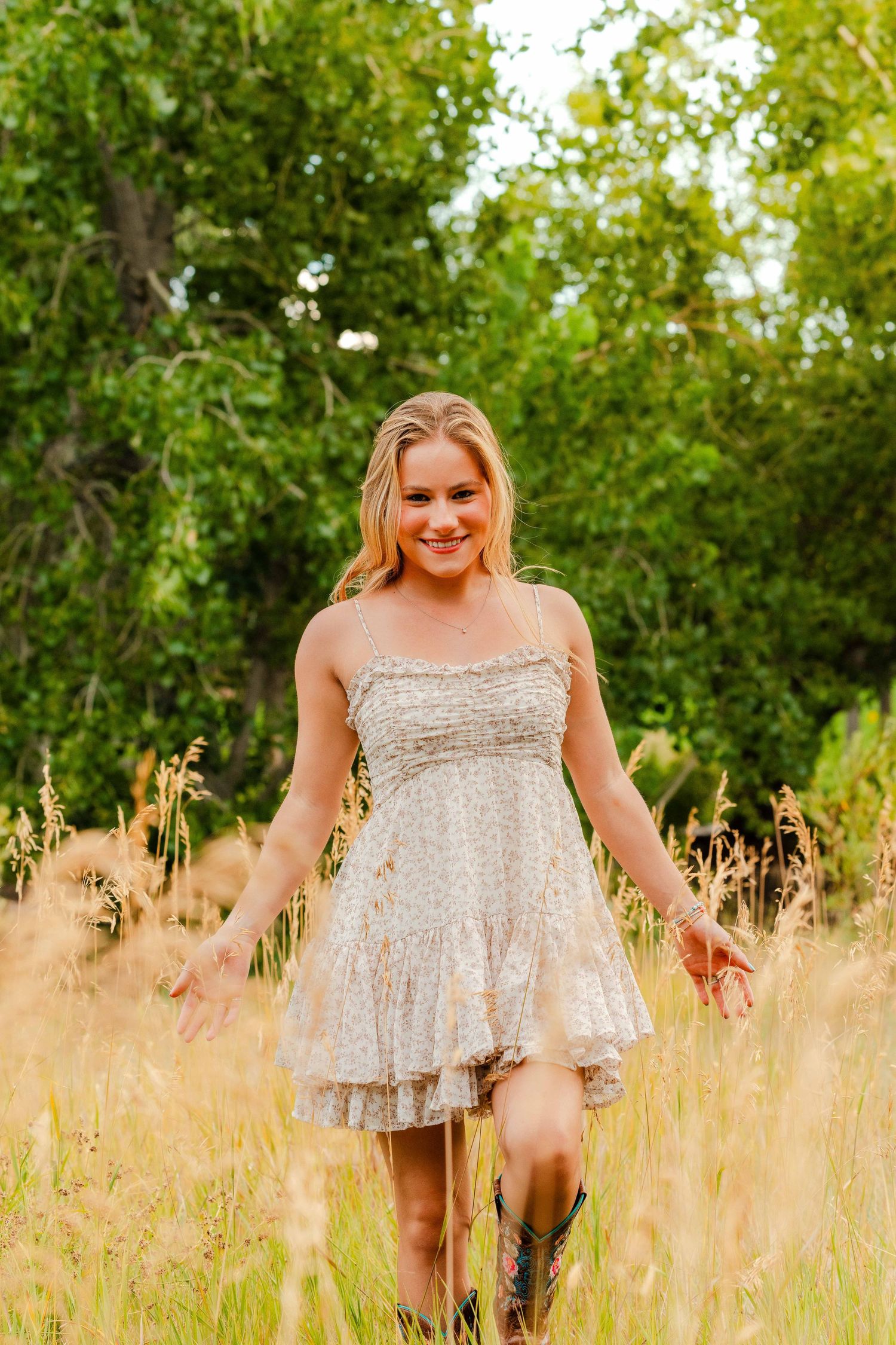 Smiling blonde woman in a floral sundress and cowboy boots walks through golden tall grass outdoors.