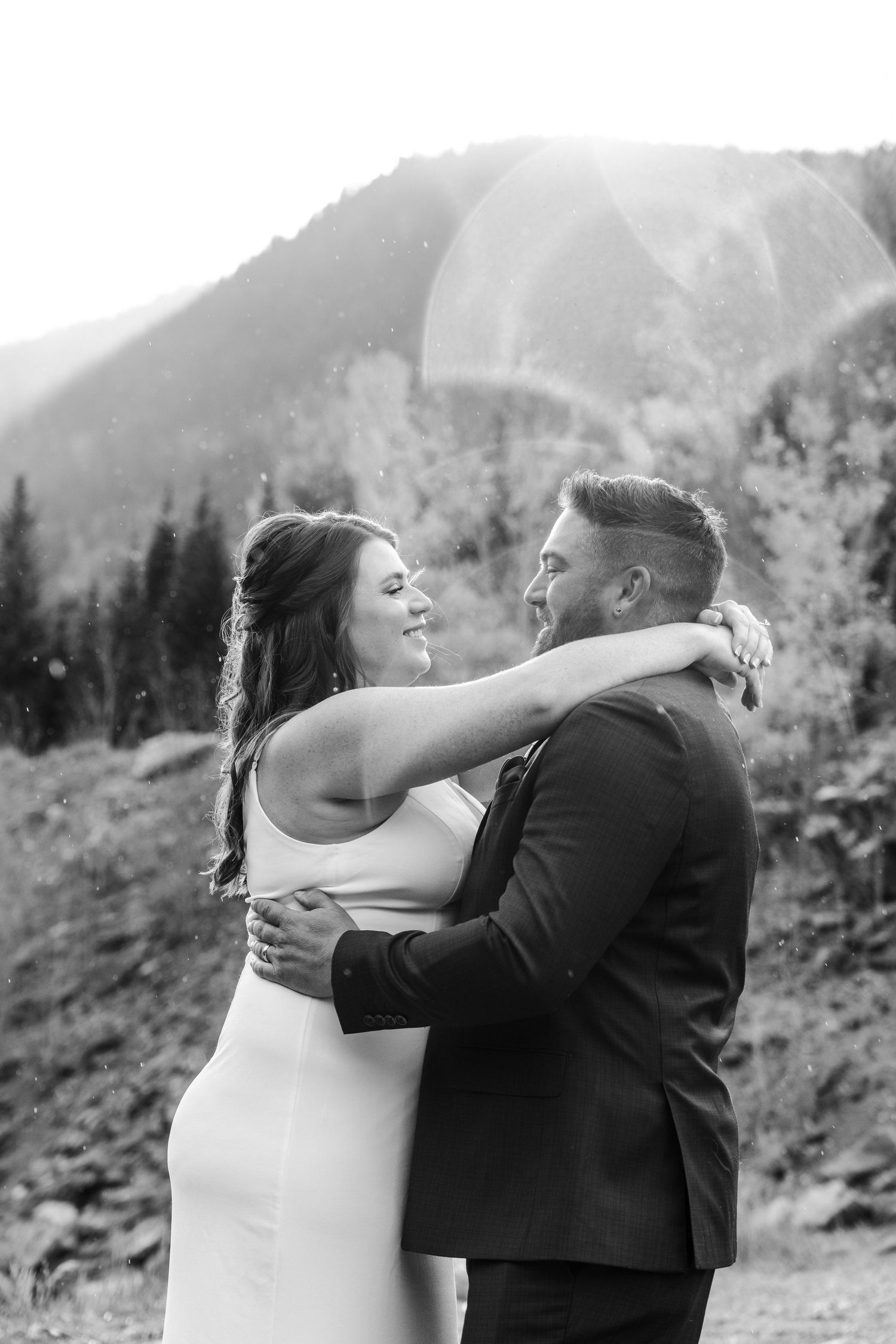 Black and white photo of bride and groom embracing with mountain forest backdrop.
