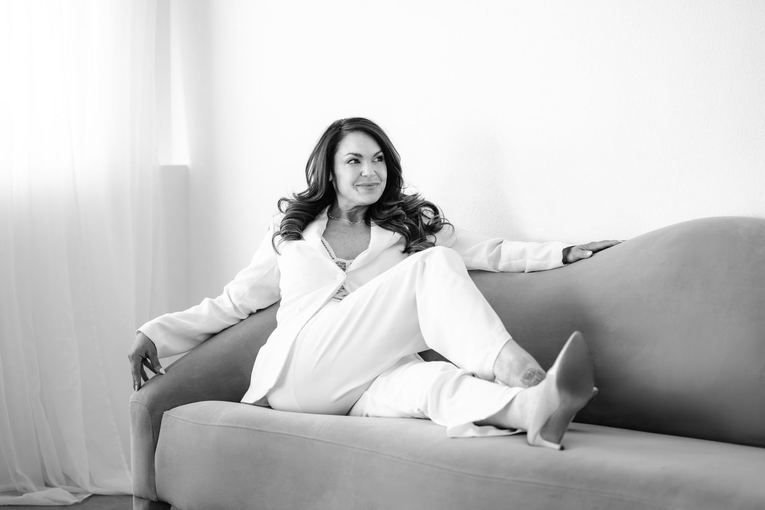 Elegant woman in white suit reclines gracefully on a sofa in black and white portrait.
