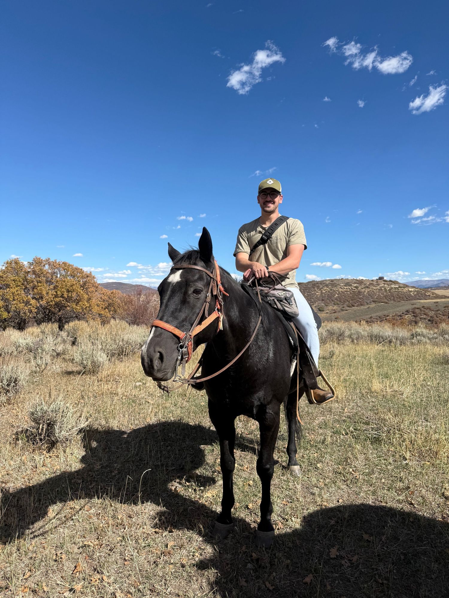 Man on dark horse in rural landscape with blue sky, wearing cap and casual clothes, riding through dry grasslands.