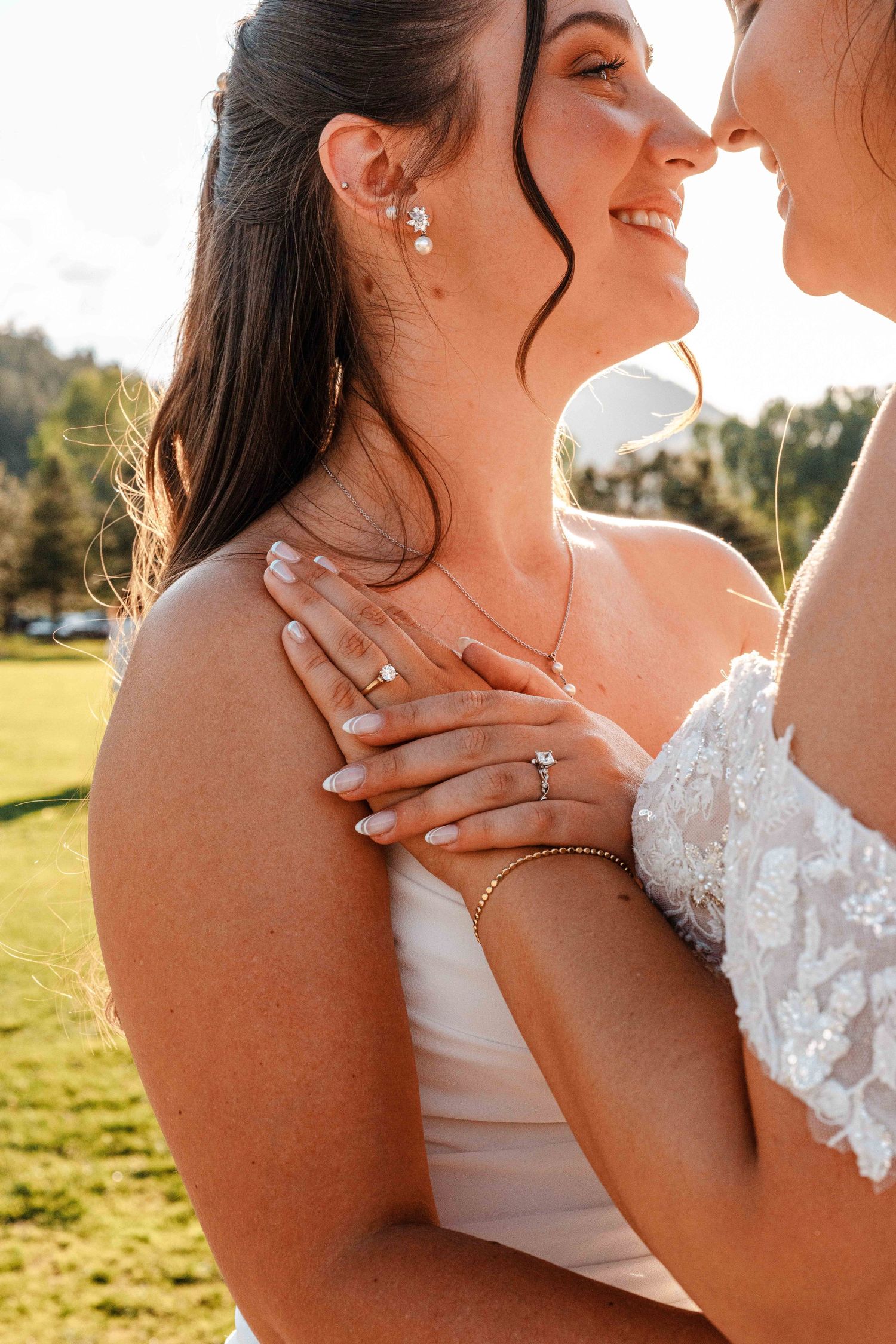 Two brides in white gowns share a tender moment, showcasing elegant jewelry and manicured nails.