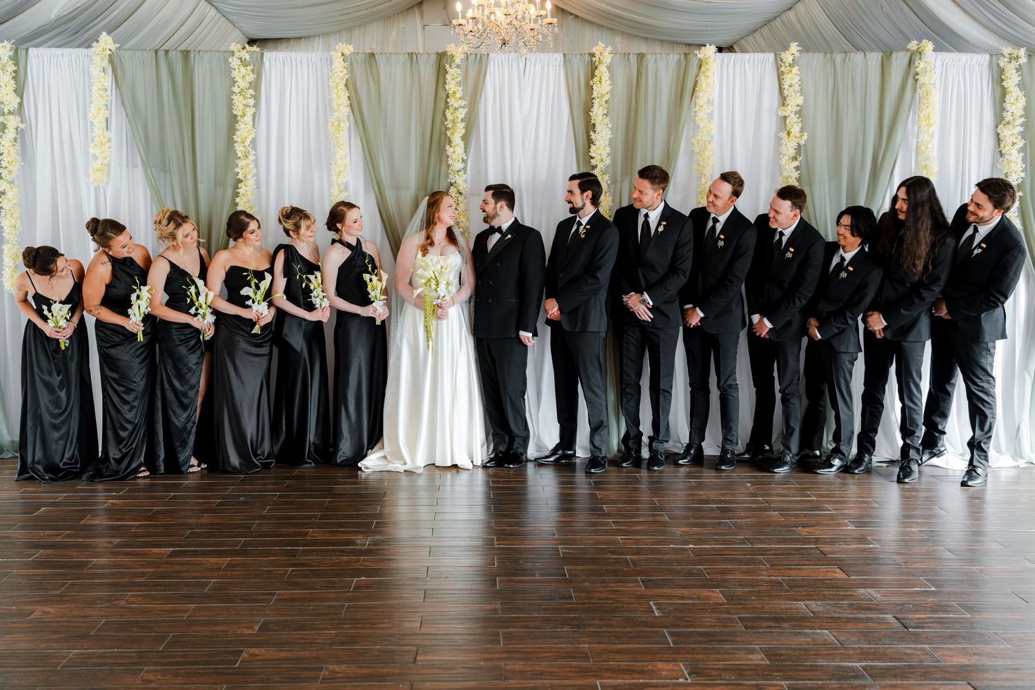 Wedding party in elegant venue, bridesmaids in black gowns and groomsmen in suits flanking bride and groom.