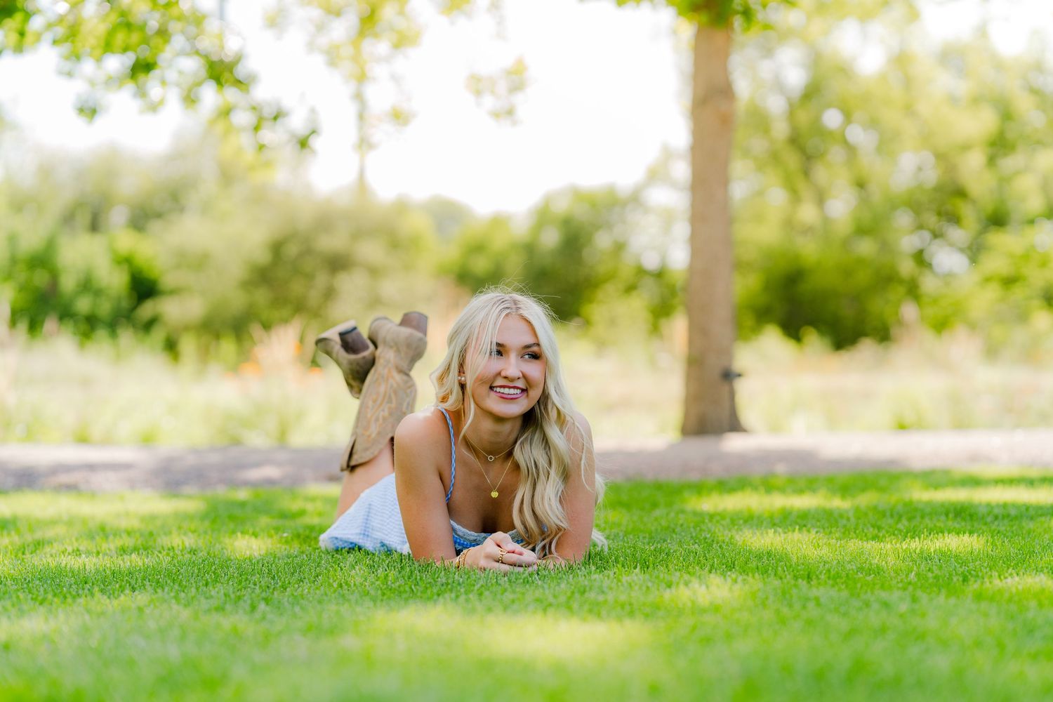 Smiling blonde woman lying on lush green grass in a sunny park, wearing a white top, with trees in the background.