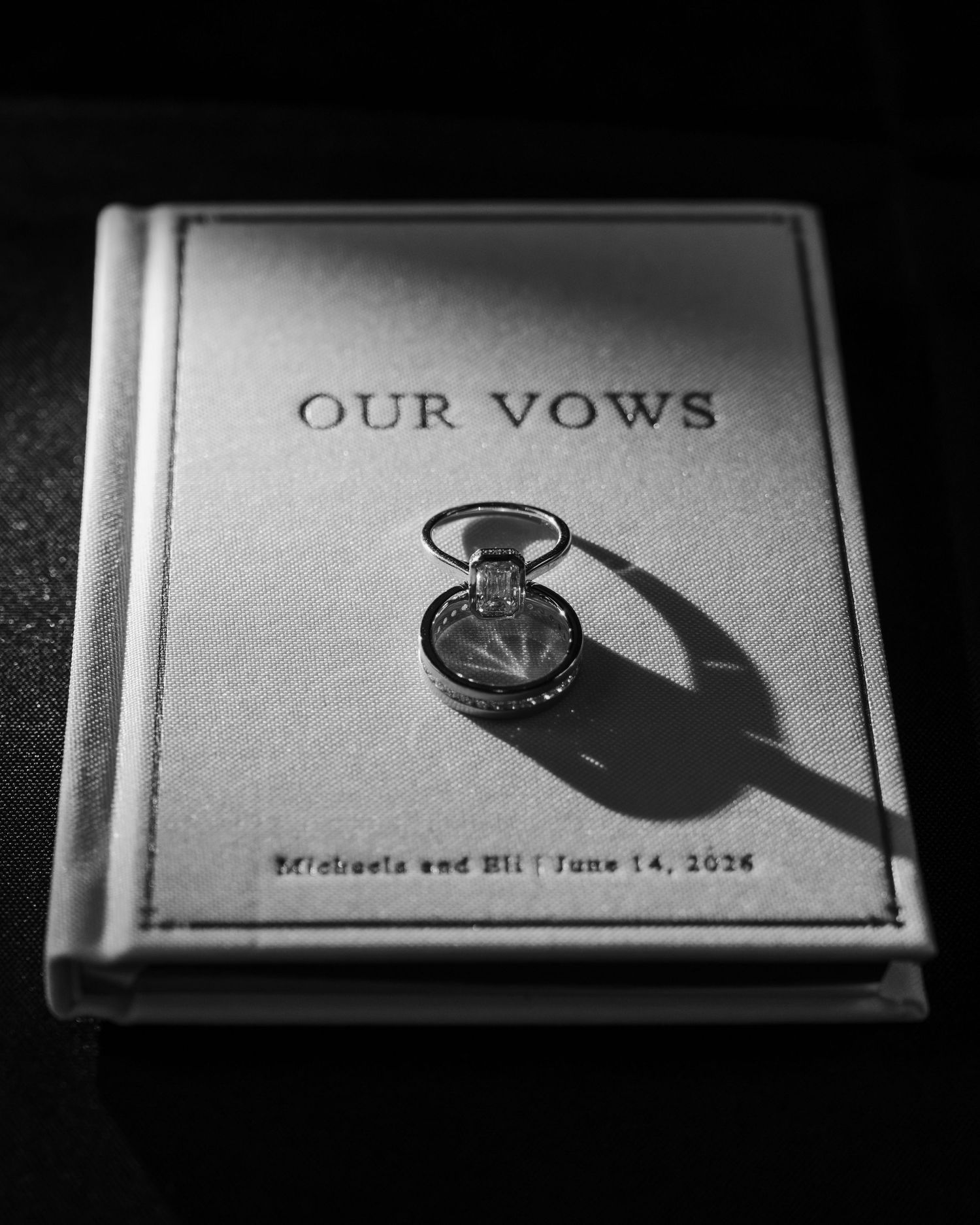 Black and white photo of wedding rings on a vow book reading Our Vows, Michaels and Eli, June 14, 2026.