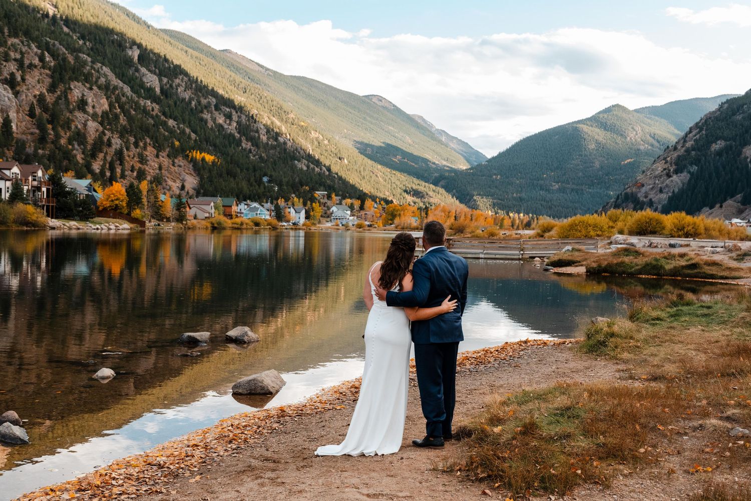 Bride and groom embrace while overlooking scenic mountain lake surrounded by autumn foliage.