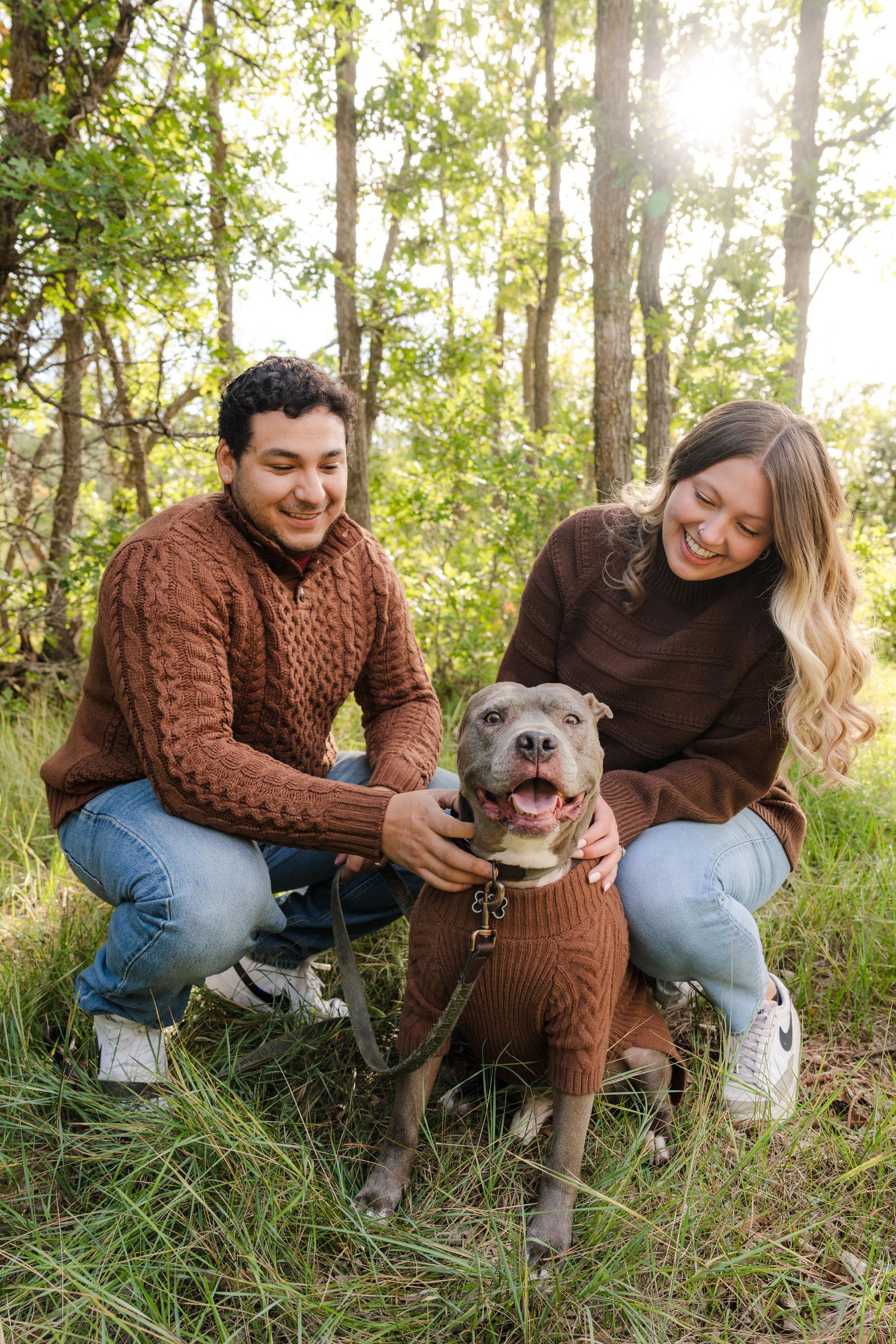 A smiling couple in matching brown sweaters crouches with their pit bull terrier wearing a sweater in a sunlit forest.
