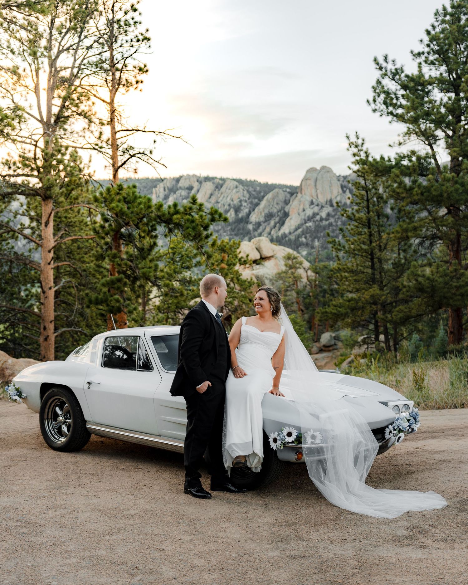 Bride and groom pose beside a vintage white Corvette with rocky mountain backdrop at sunset wedding.