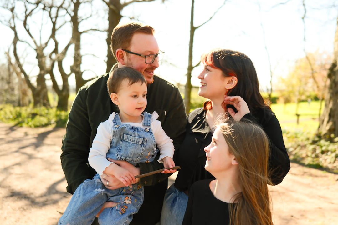 A family shares a joyful moment together on a sunny path surrounded by bare trees in a natural setting.