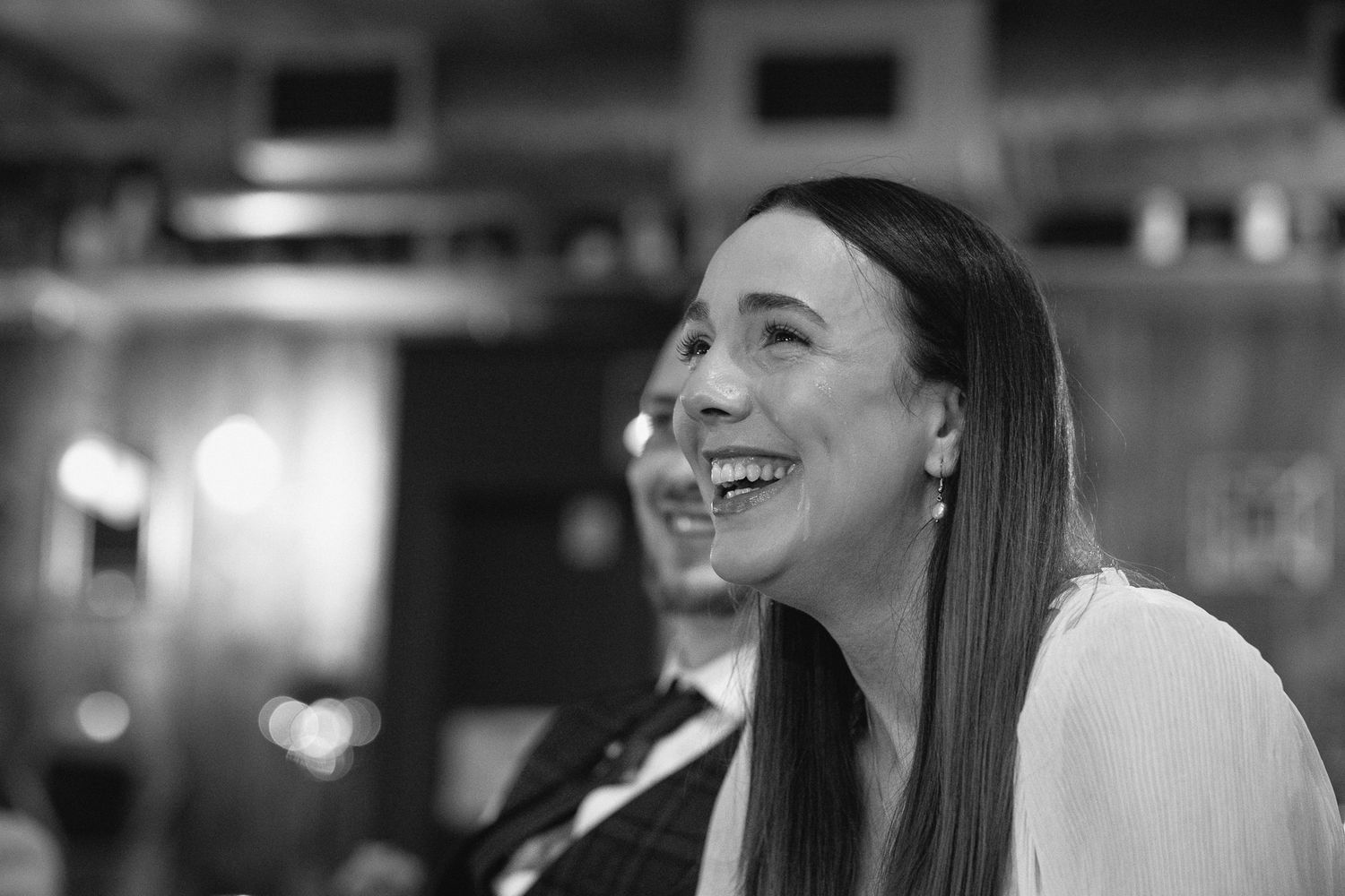 Black and white photo of a smiling woman with long hair laughing joyfully at an indoor event.