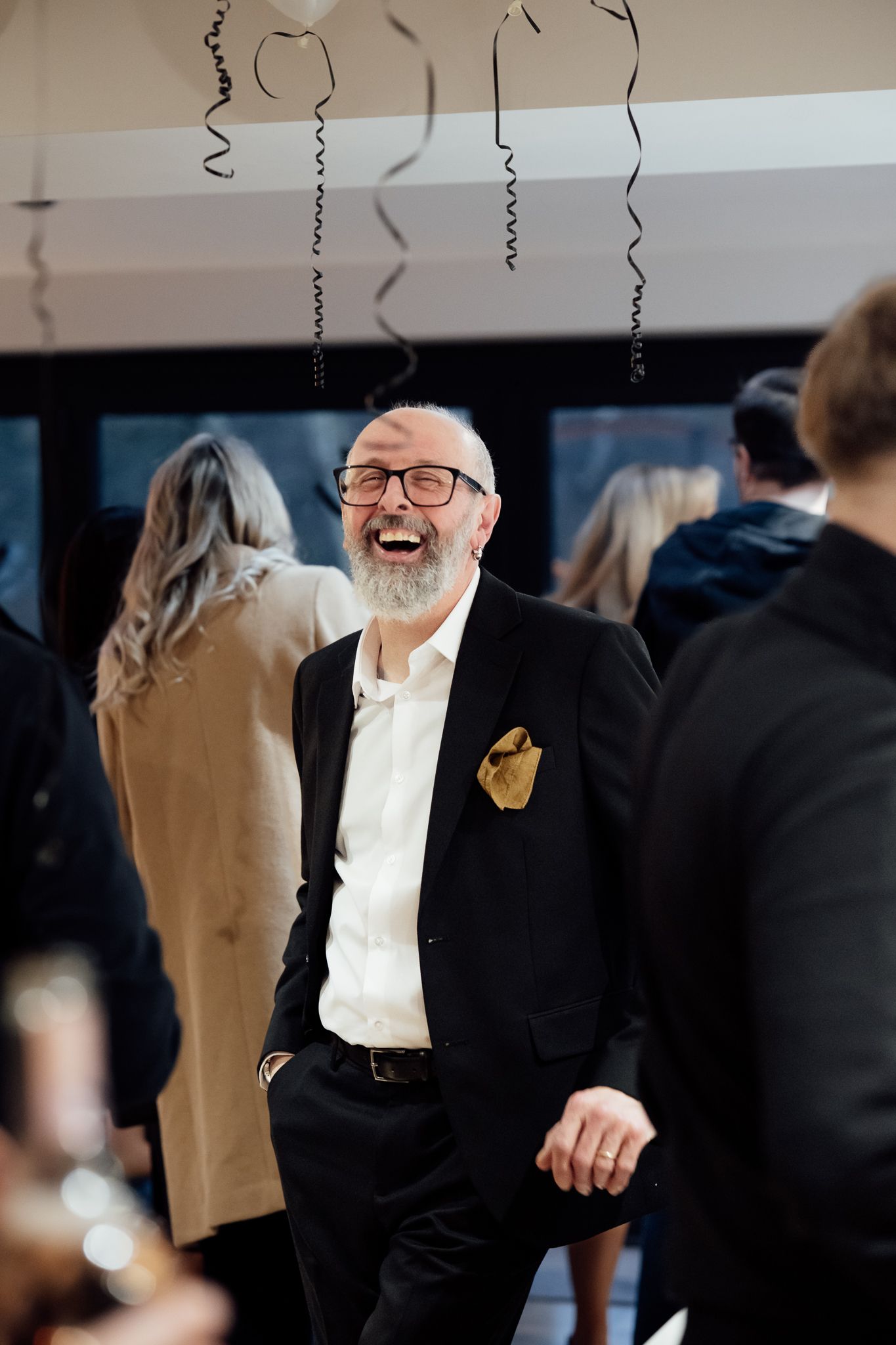 Laughing older man with glasses and beard wearing black suit with gold pocket square at festive party.