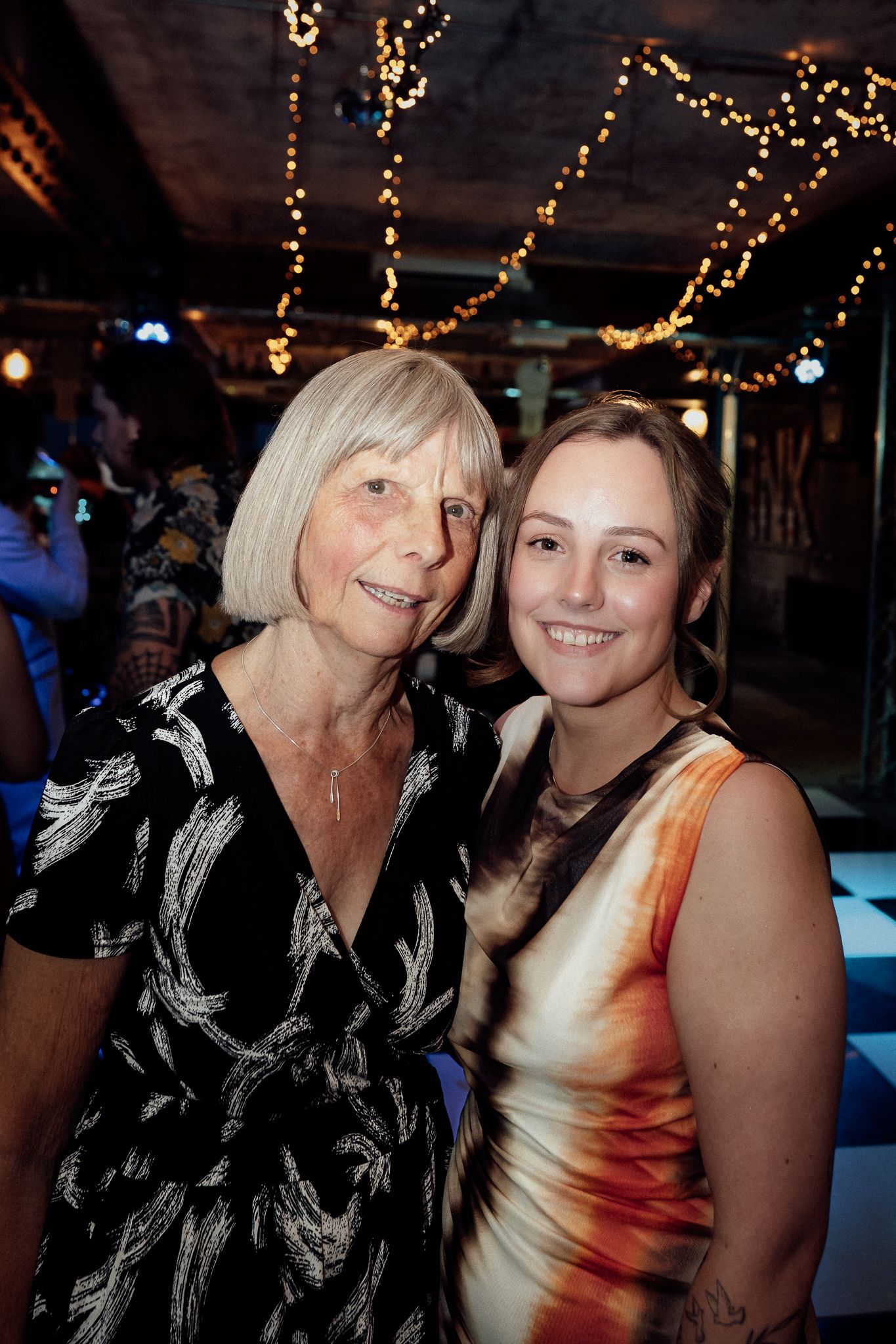 Two women smiling together at an indoor event decorated with string lights in the background.