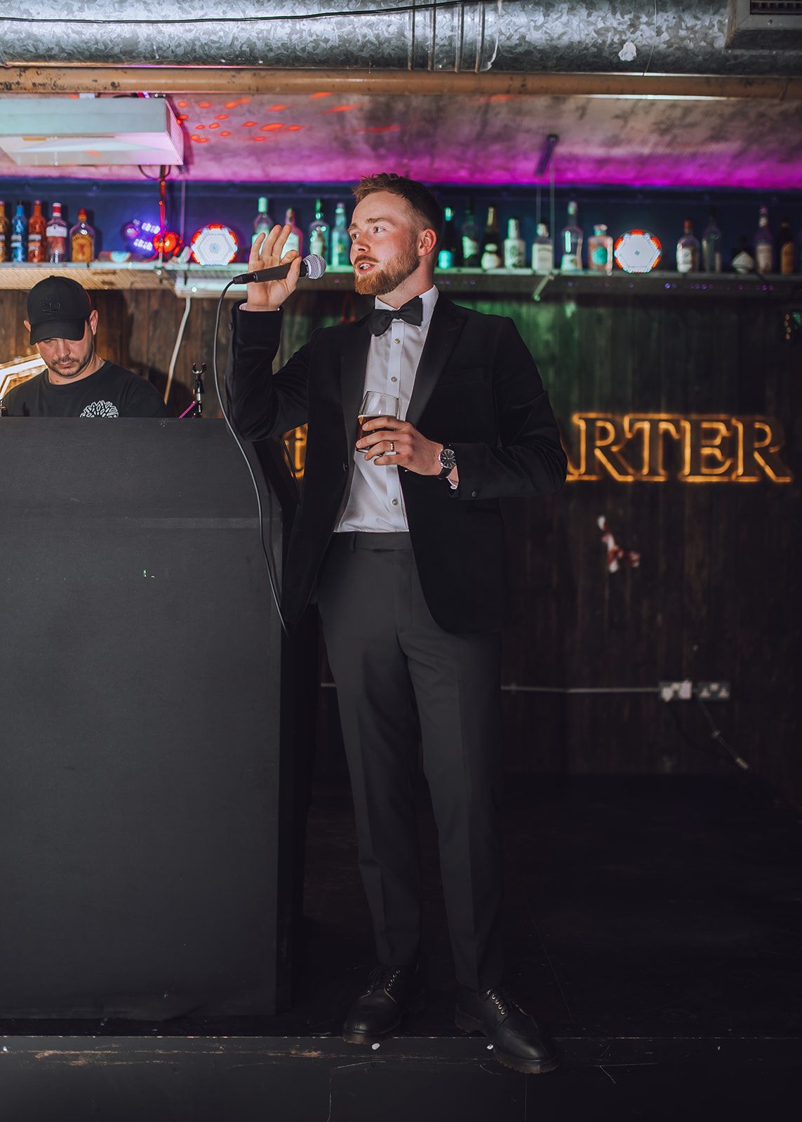 A bartender in formal attire pours drinks behind a dimly lit bar counter with bottles visible in the background.