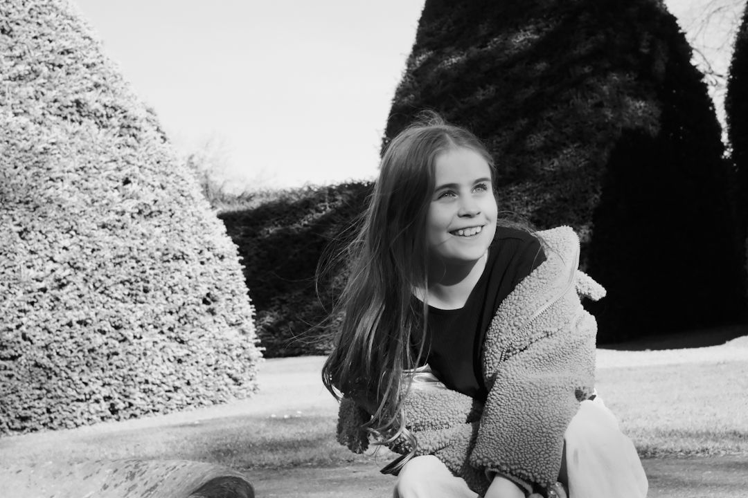 Black and white photo of a smiling girl with long hair sitting outdoors near trimmed hedges.
