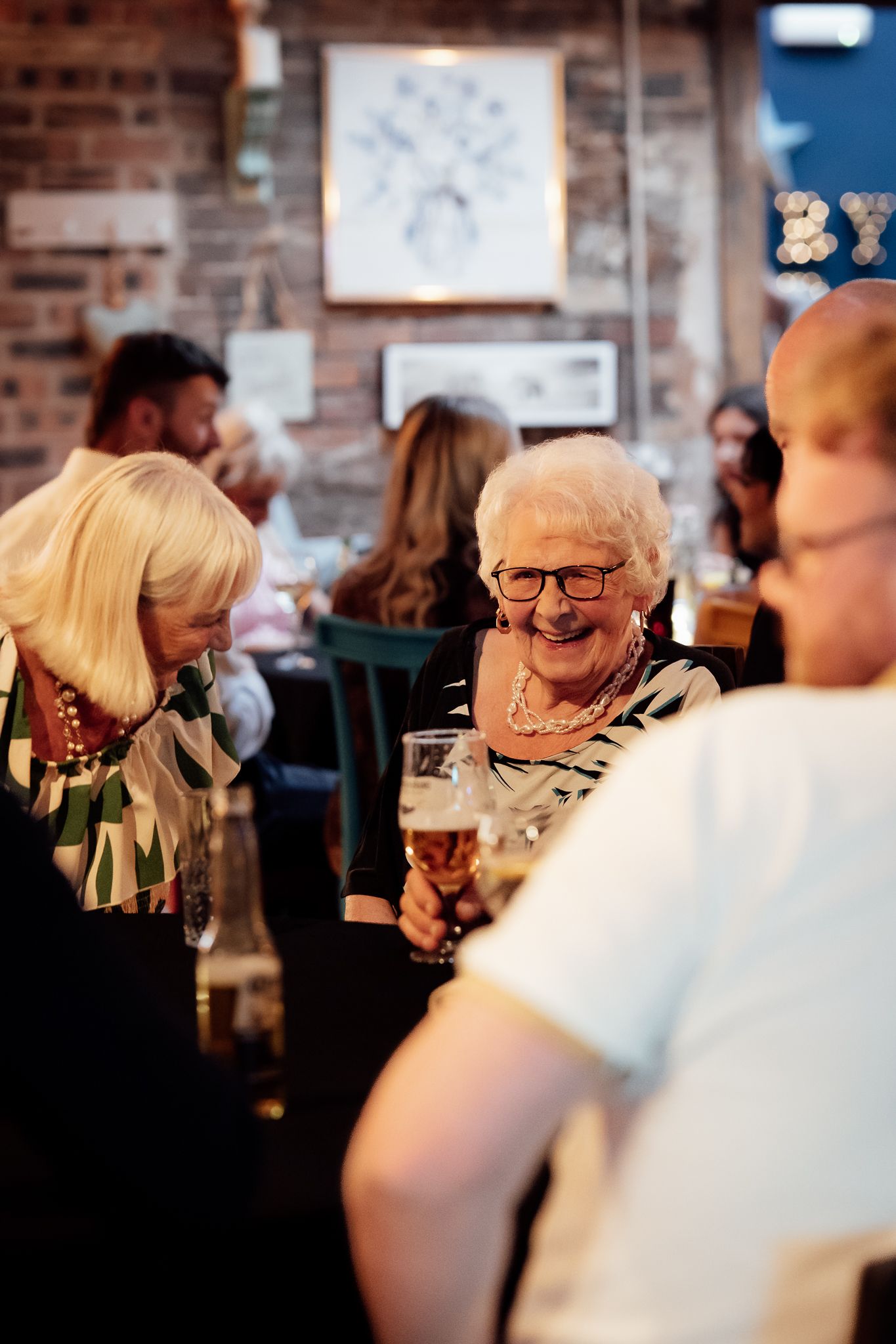 Elderly woman laughing and holding a beer glass at a lively pub gathering with friends.