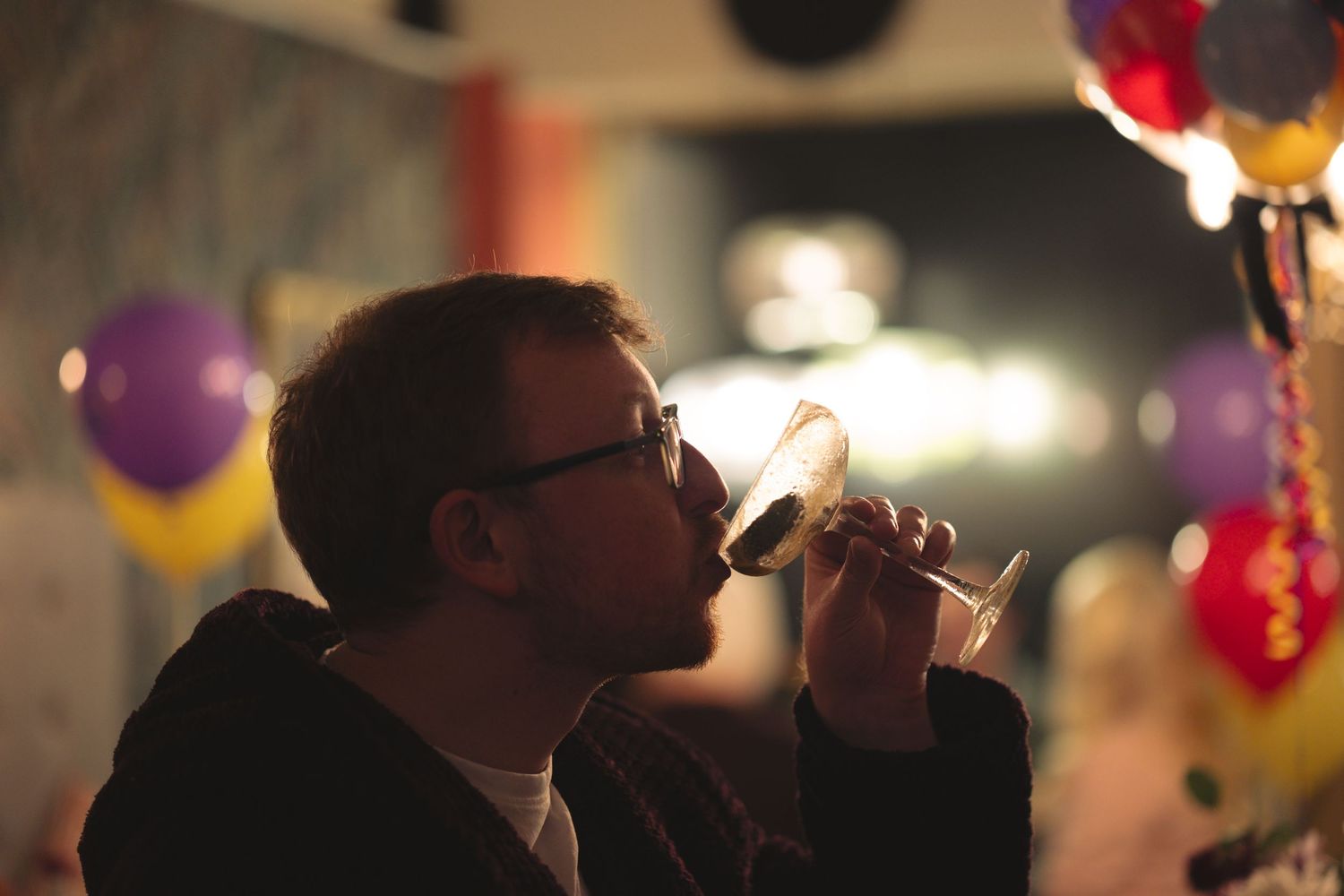 A person in glasses sips from a wine glass at a party decorated with colorful balloons in the background.