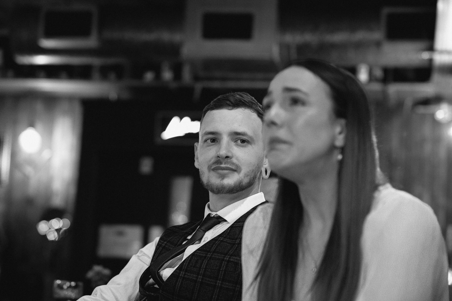 Black and white photo of a man in a vest and woman with long hair at an indoor event.