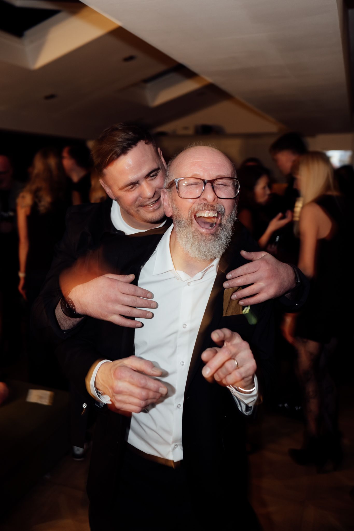 People hugging and laughing together at a dimly lit indoor celebration.