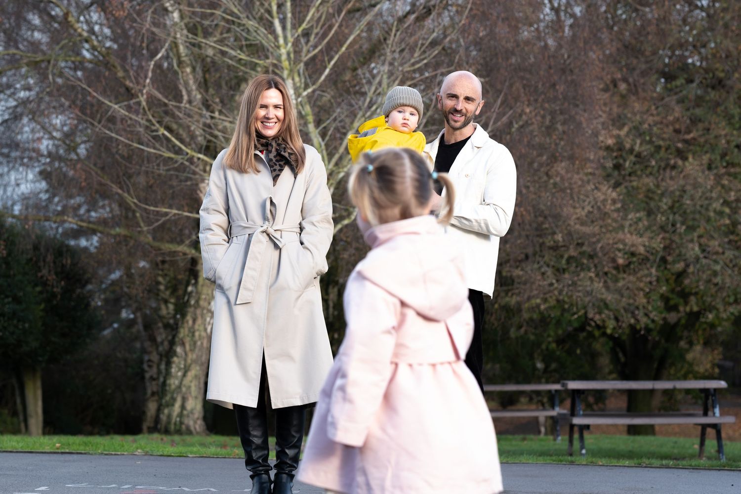 A family in winter coats poses together outdoors on a path with bare trees in the background.