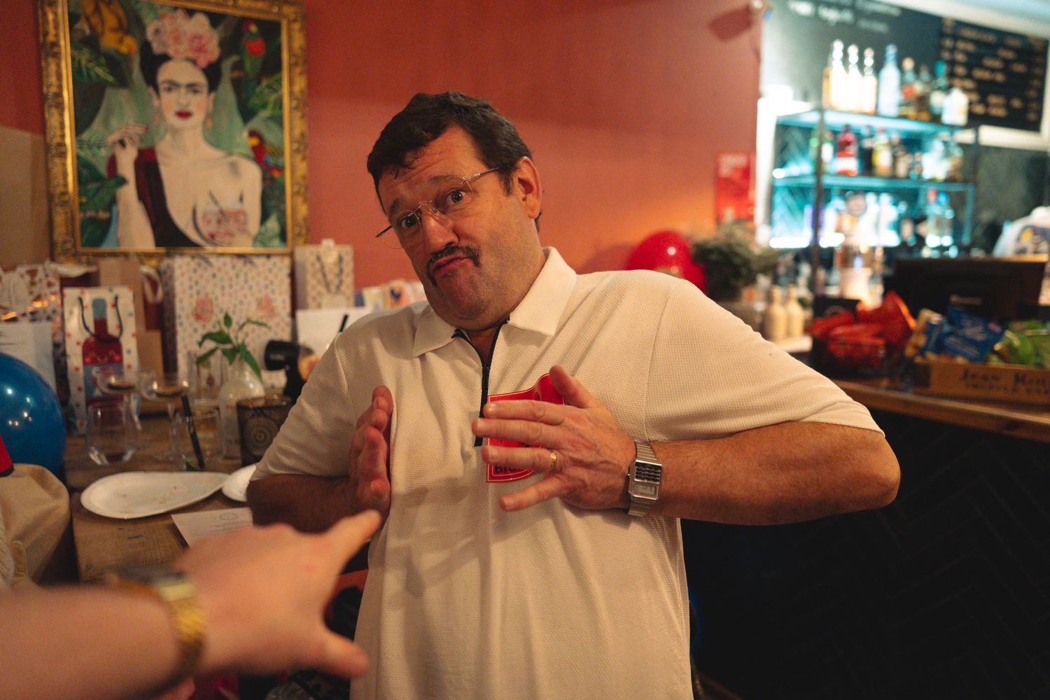 Several people sitting at a bar counter in a casual setting with red walls and bottles in the background.