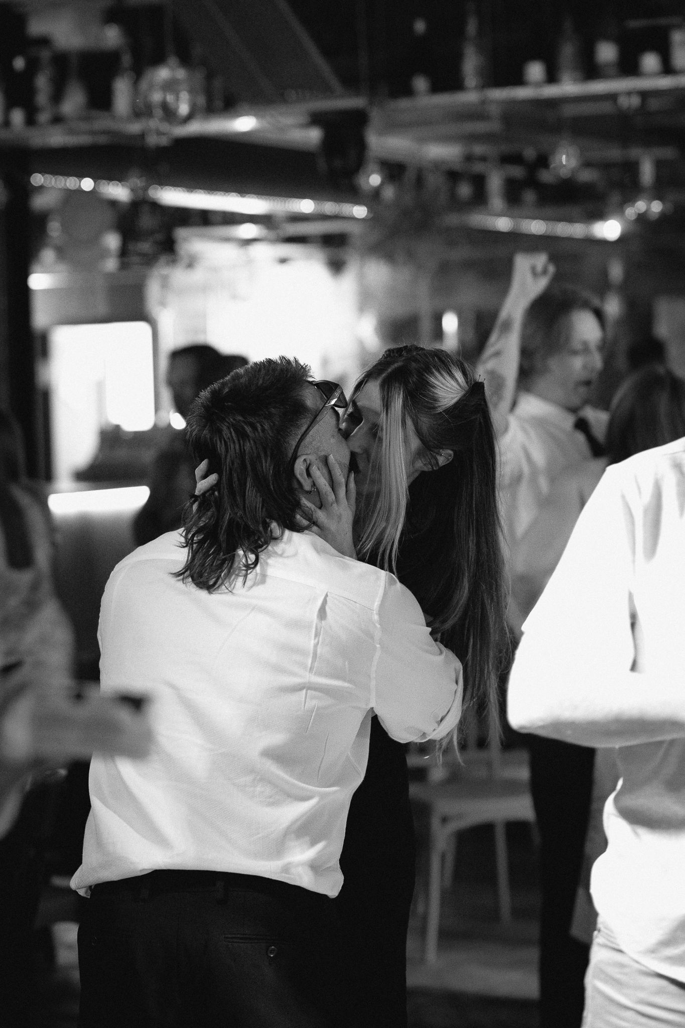 Black and white photo of a couple kissing on the dance floor at a lively indoor party.