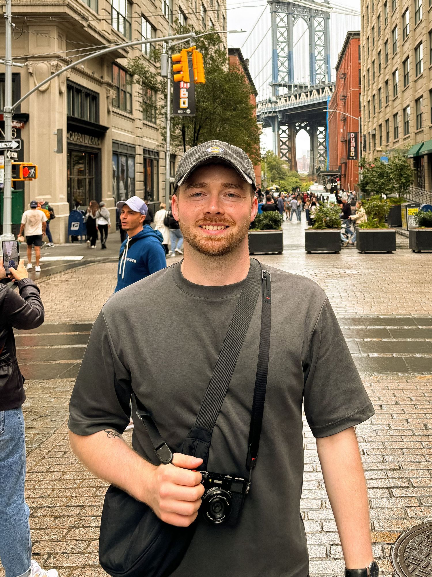 Person in gray t-shirt and baseball cap stands on cobblestone street with Manhattan Bridge visible in background.