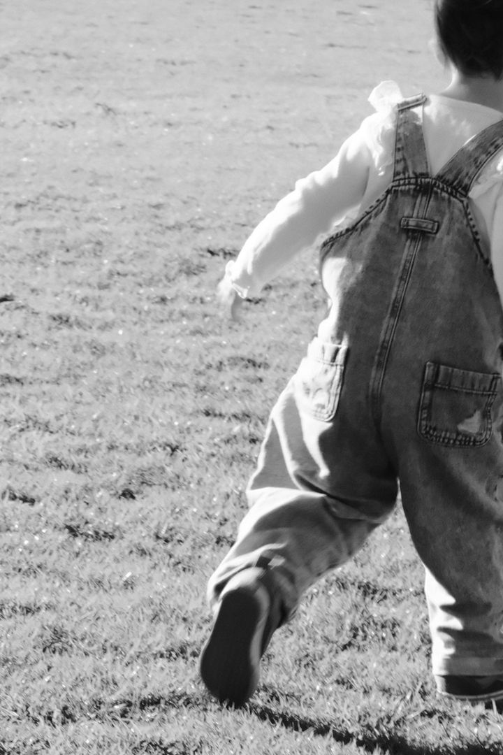 Black and white photo of a child in overalls walking across a grassy field in sunlight.