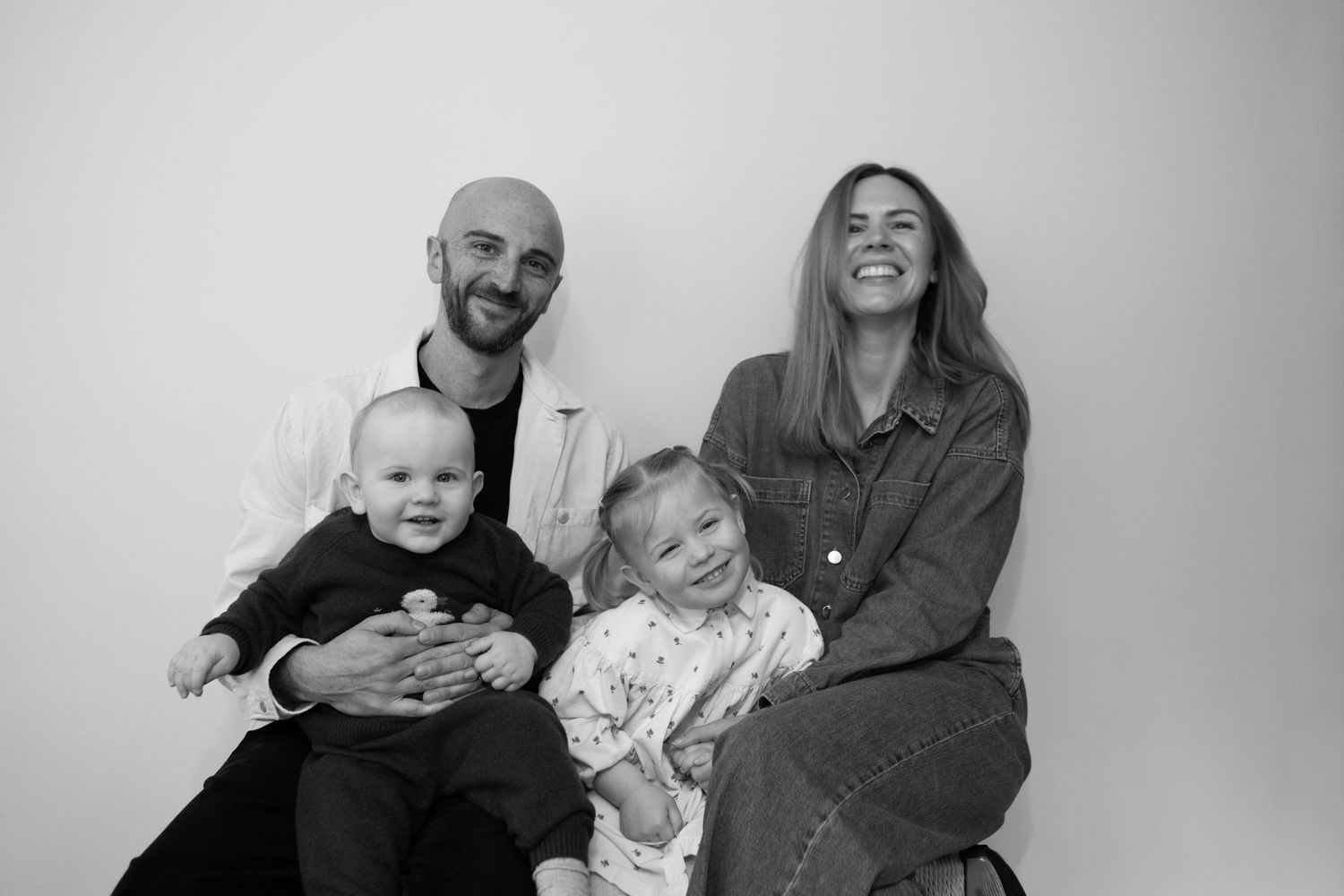 A family of four poses together for a casual black and white portrait against a plain background.