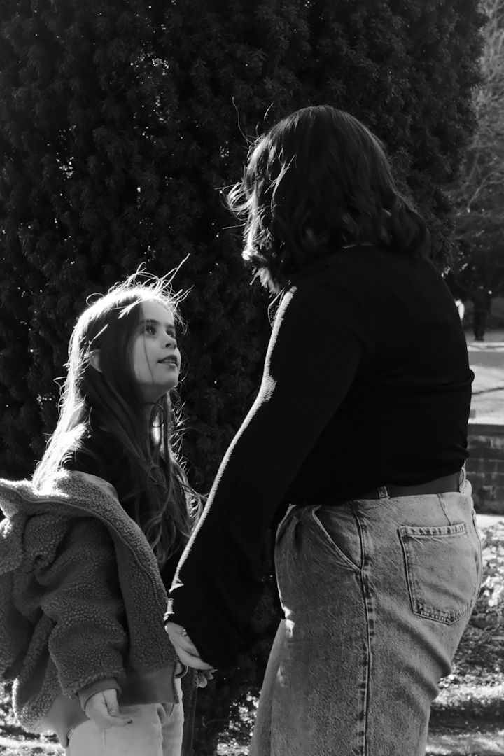 A black and white candid photo of two people in casual attire having a conversation outdoors near evergreen trees.