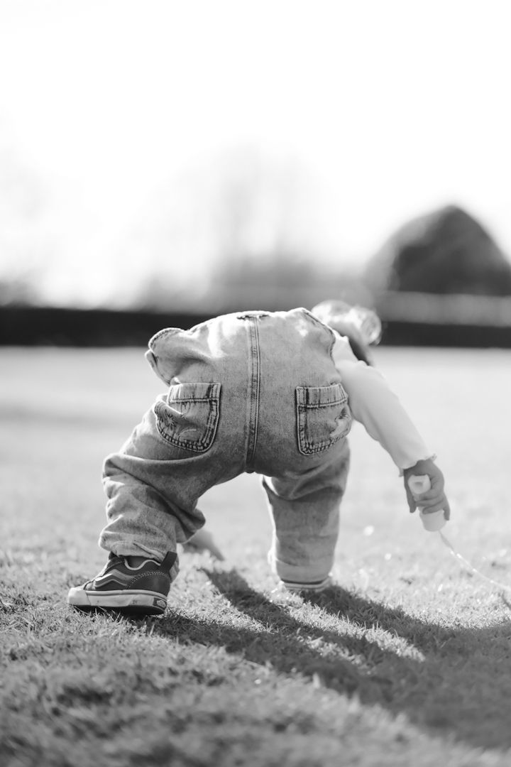 Black and white photo of a small child in denim overalls bending down to touch the grass in soft sunlight.