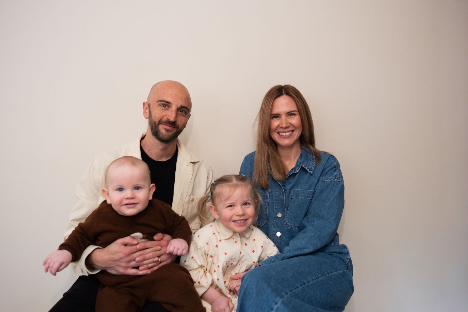 A smiling family of four with a bald father, mother in denim, and two young children posing together against a white wall.