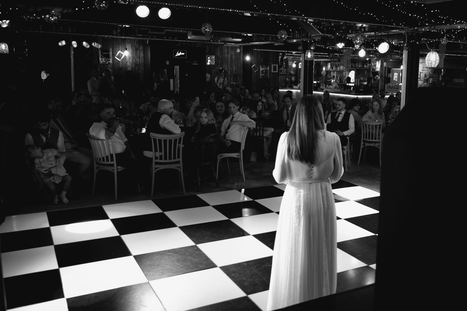 Bride stands on black and white checkered dance floor facing seated wedding guests in elegant venue.