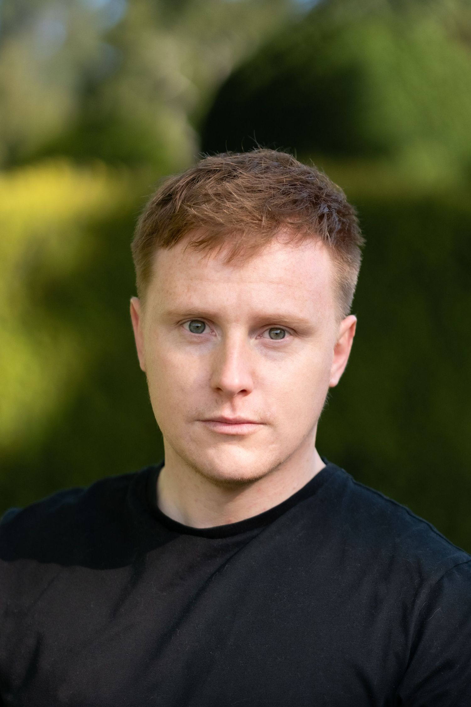 Portrait photo of a person wearing a black t-shirt against a blurred green outdoor background.